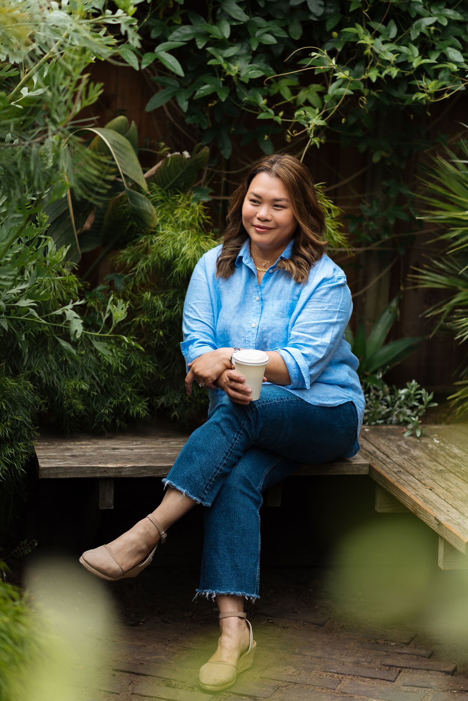 Person in denim outfit sitting on wooden deck surrounded by lush green foliage.