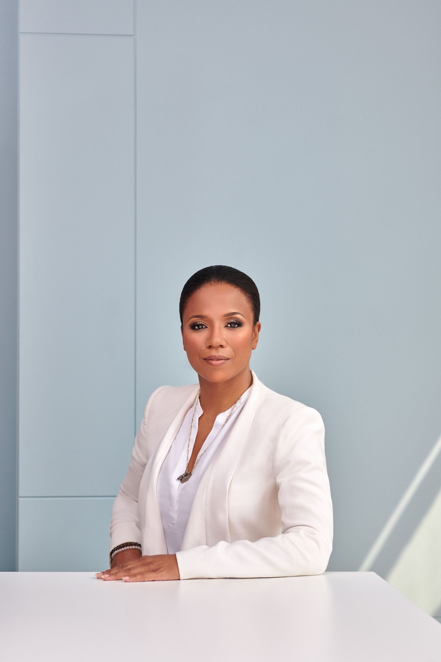 Professional Black businesswoman in white blazer sitting at white desk against light blue wall.