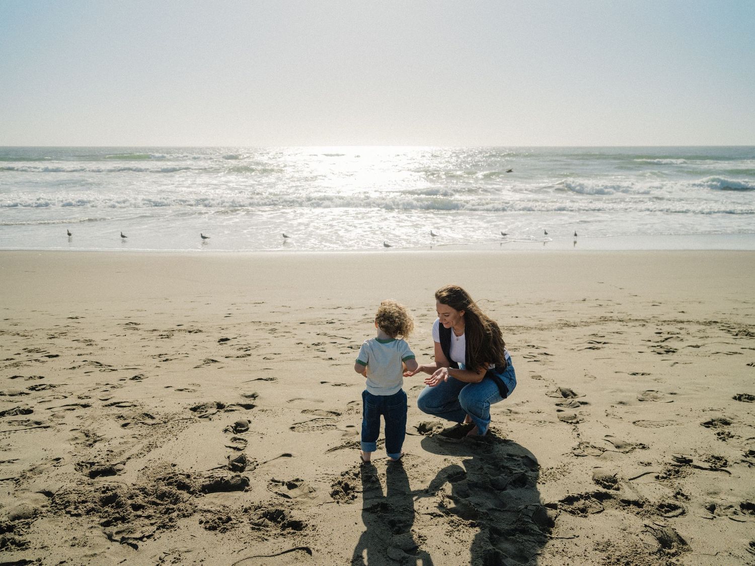 A mother kneels on a sandy beach interacting with her toddler near the ocean shoreline on a sunny day.