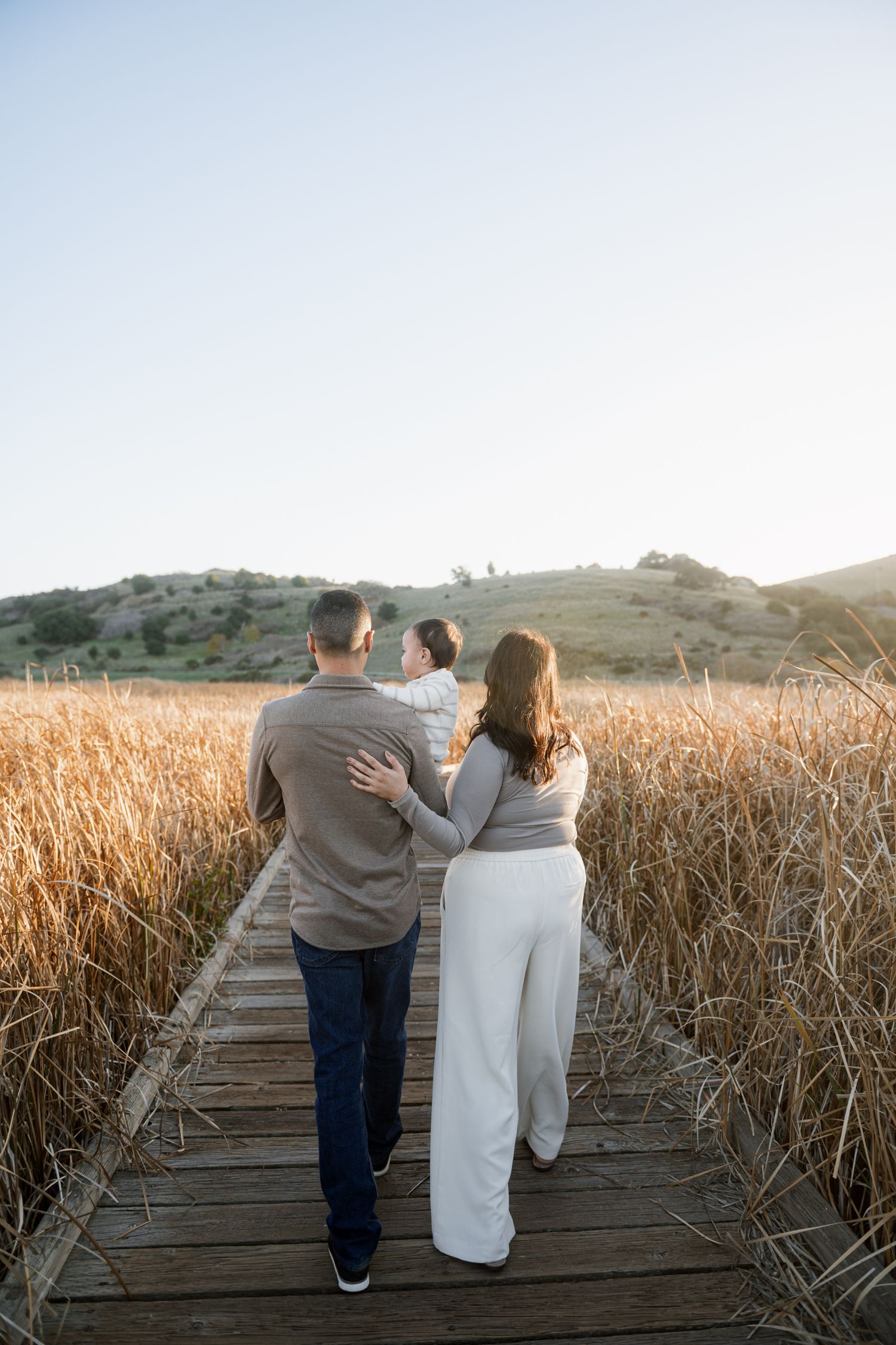 Couple walking together on a wooden boardwalk through golden wheat fields at sunset.