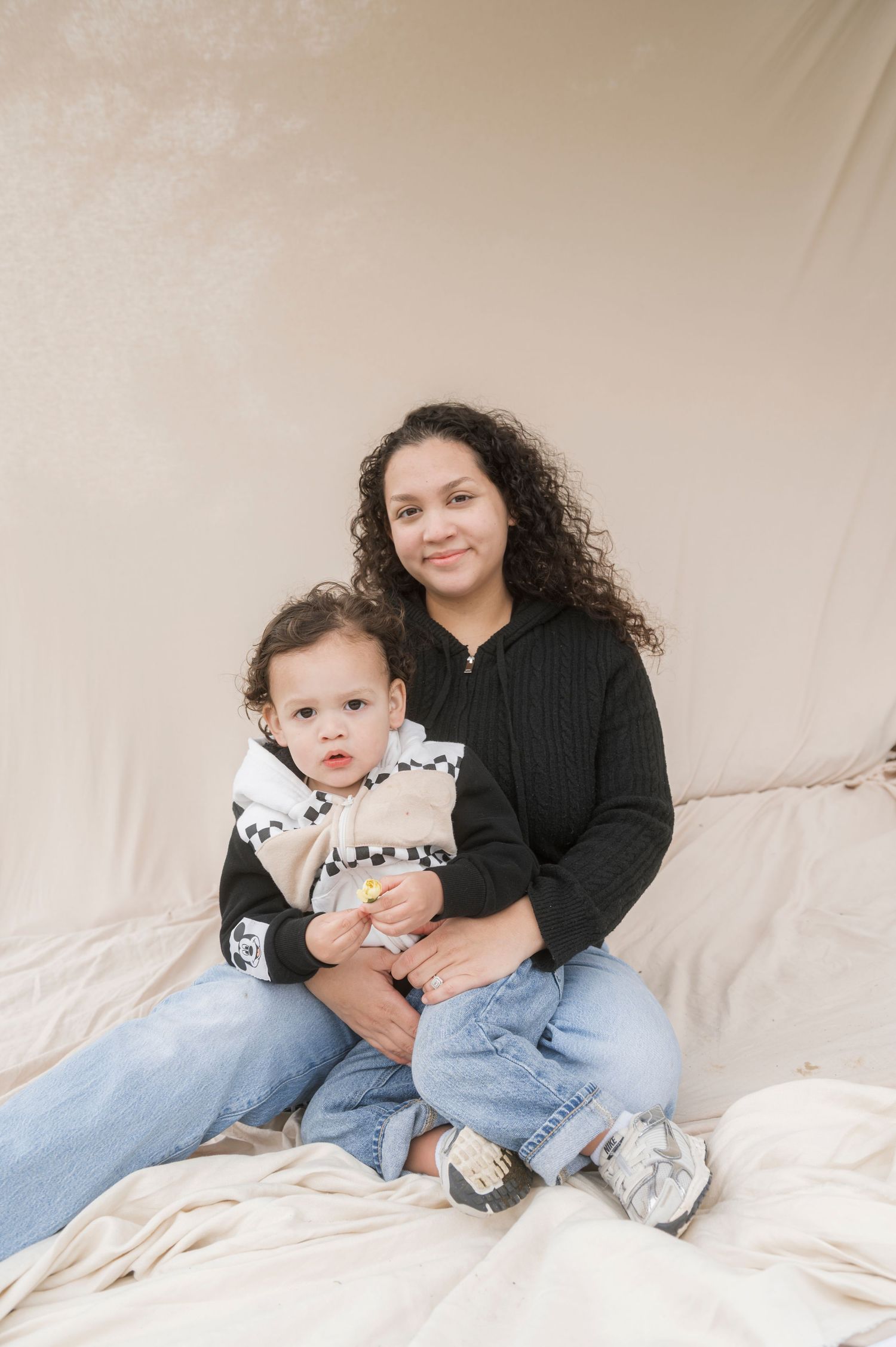 A young woman with curly hair in a black hoodie and jeans sits holding a toddler against a neutral backdrop.