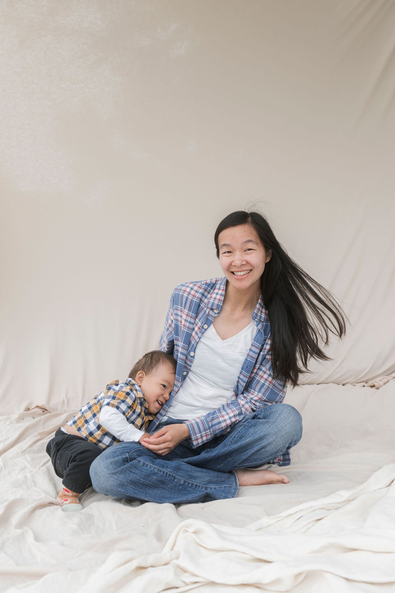 Smiling Asian woman in plaid shirt sitting cross-legged on white fabric with a toddler leaning against her.