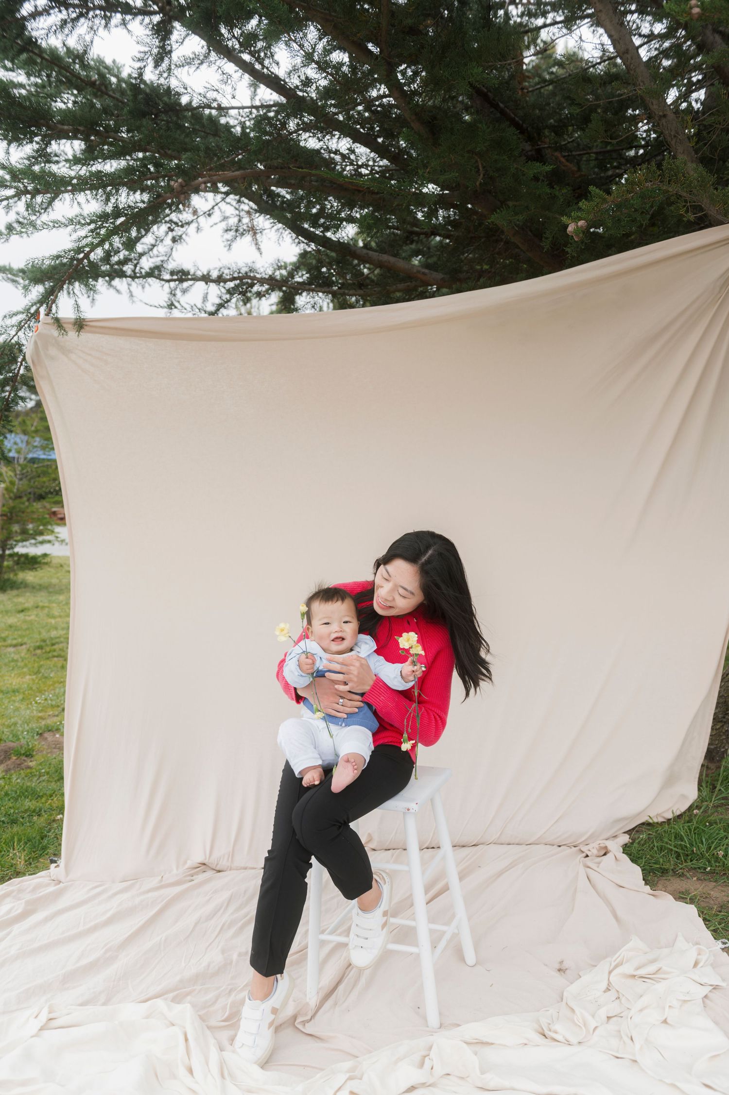 A smiling woman in a red sweater sits on a stool holding a baby outdoors against a beige fabric backdrop.