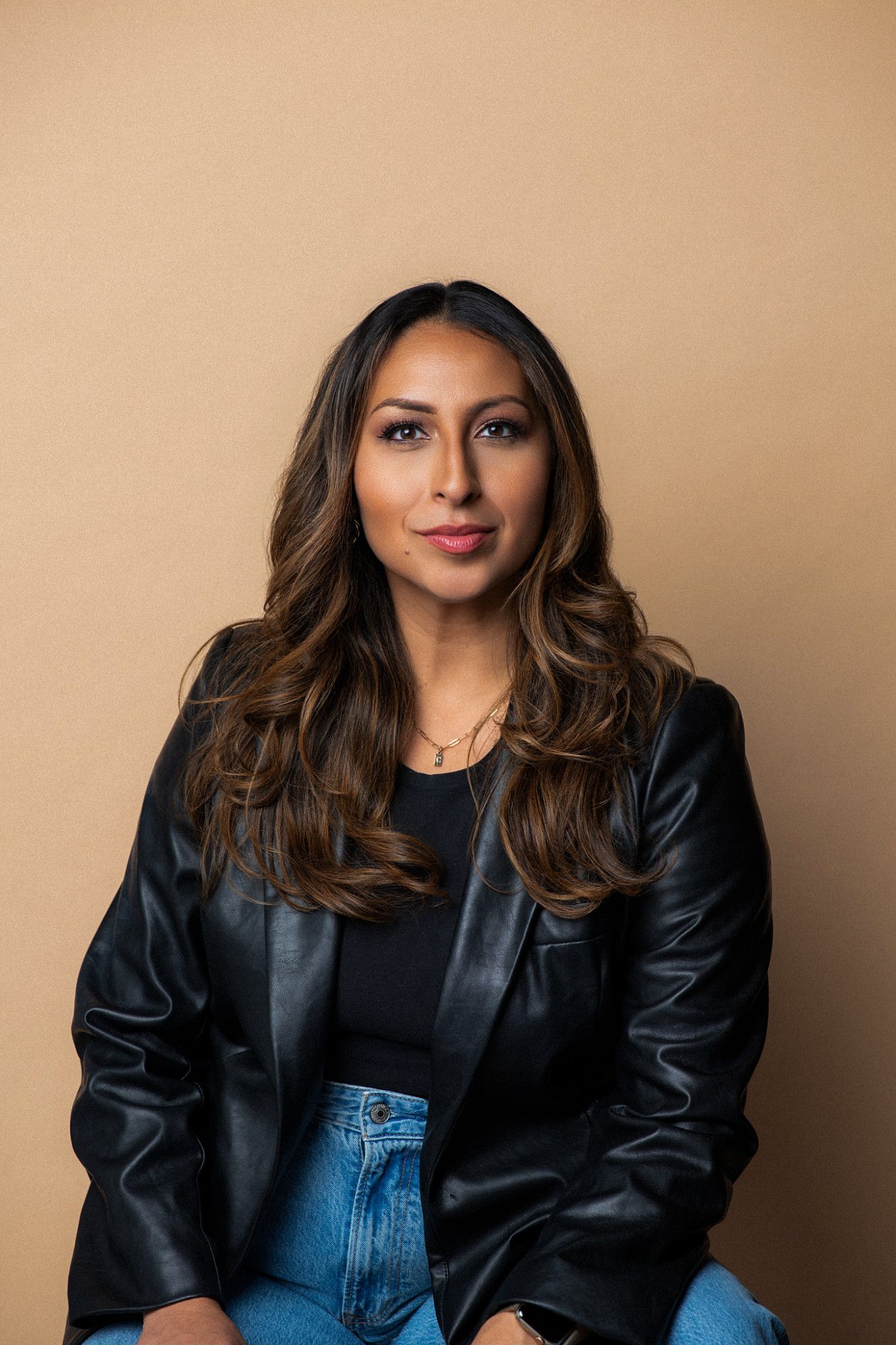 Woman in black leather blazer and jeans poses confidently against a warm beige background.