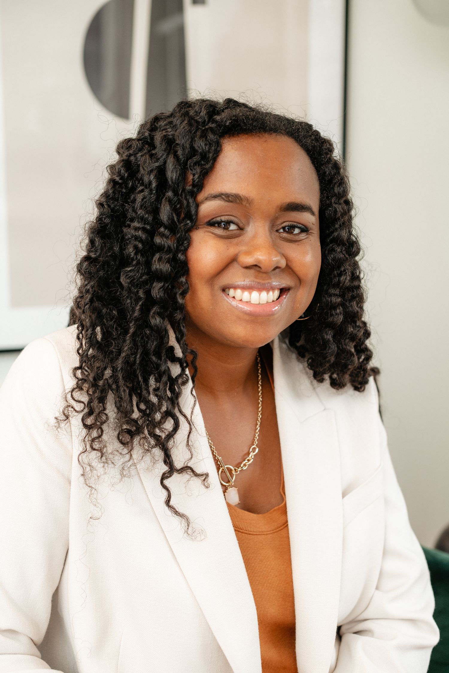 Professional portrait of someone wearing a white blazer and orange top, with curly dark hair and a bright smile.