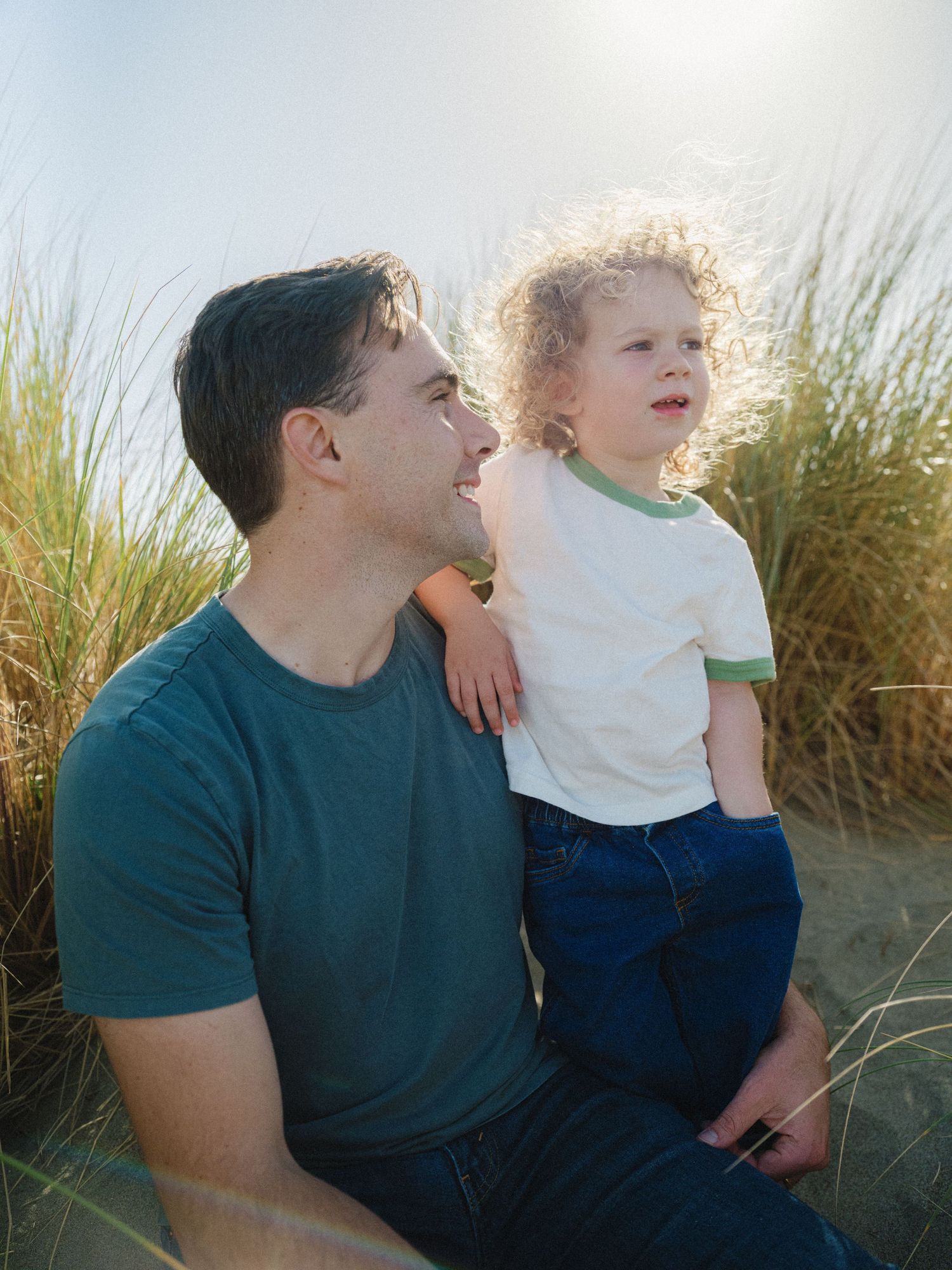 A father and young curly-haired child sit together on sandy dunes, sharing a joyful moment in warm sunlight.