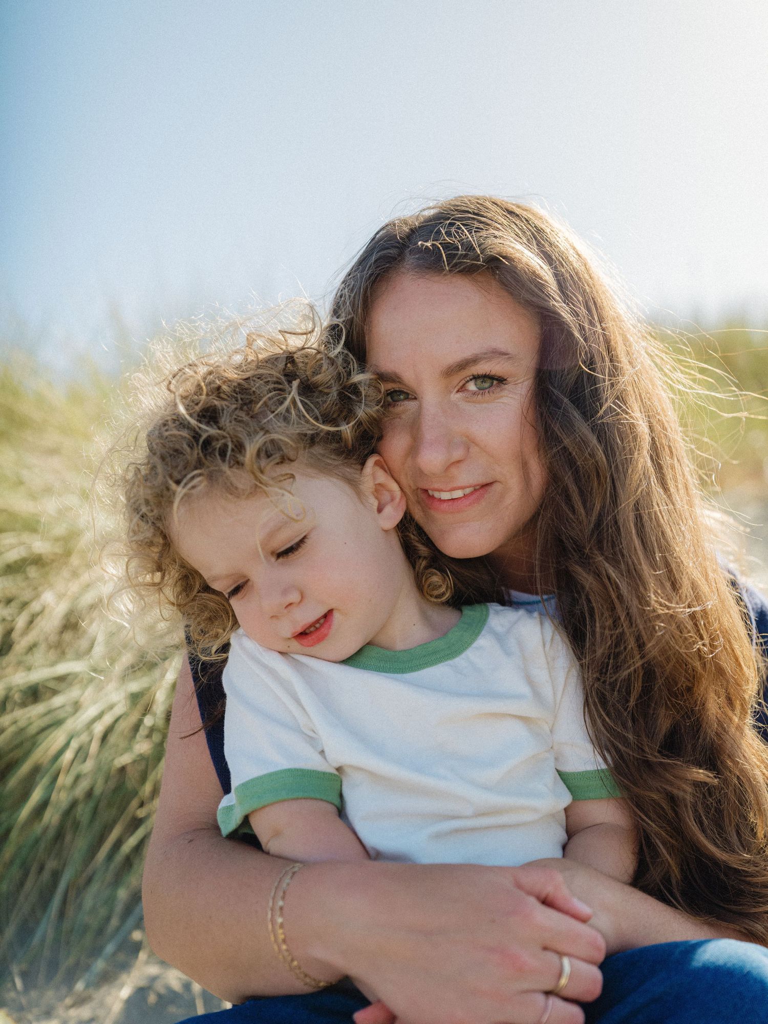 A mother with long brown hair holds her curly-haired toddler on a sunny beach, both sharing a tender moment.