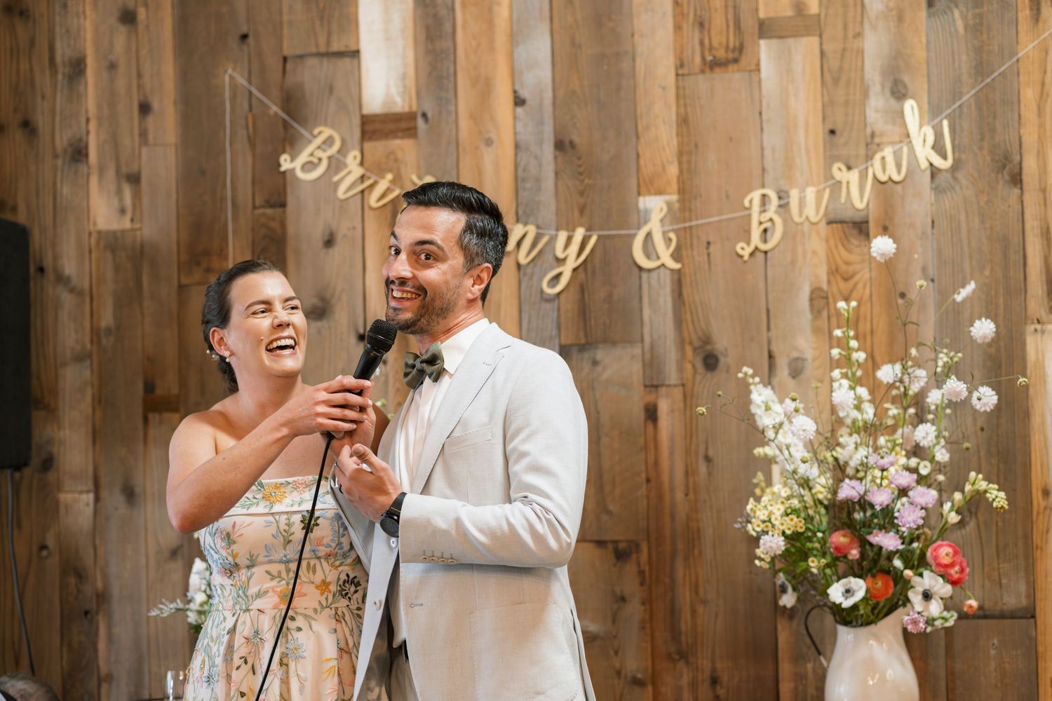 Bride and groom laughing together at microphone during wedding reception speech in rustic barn venue.
