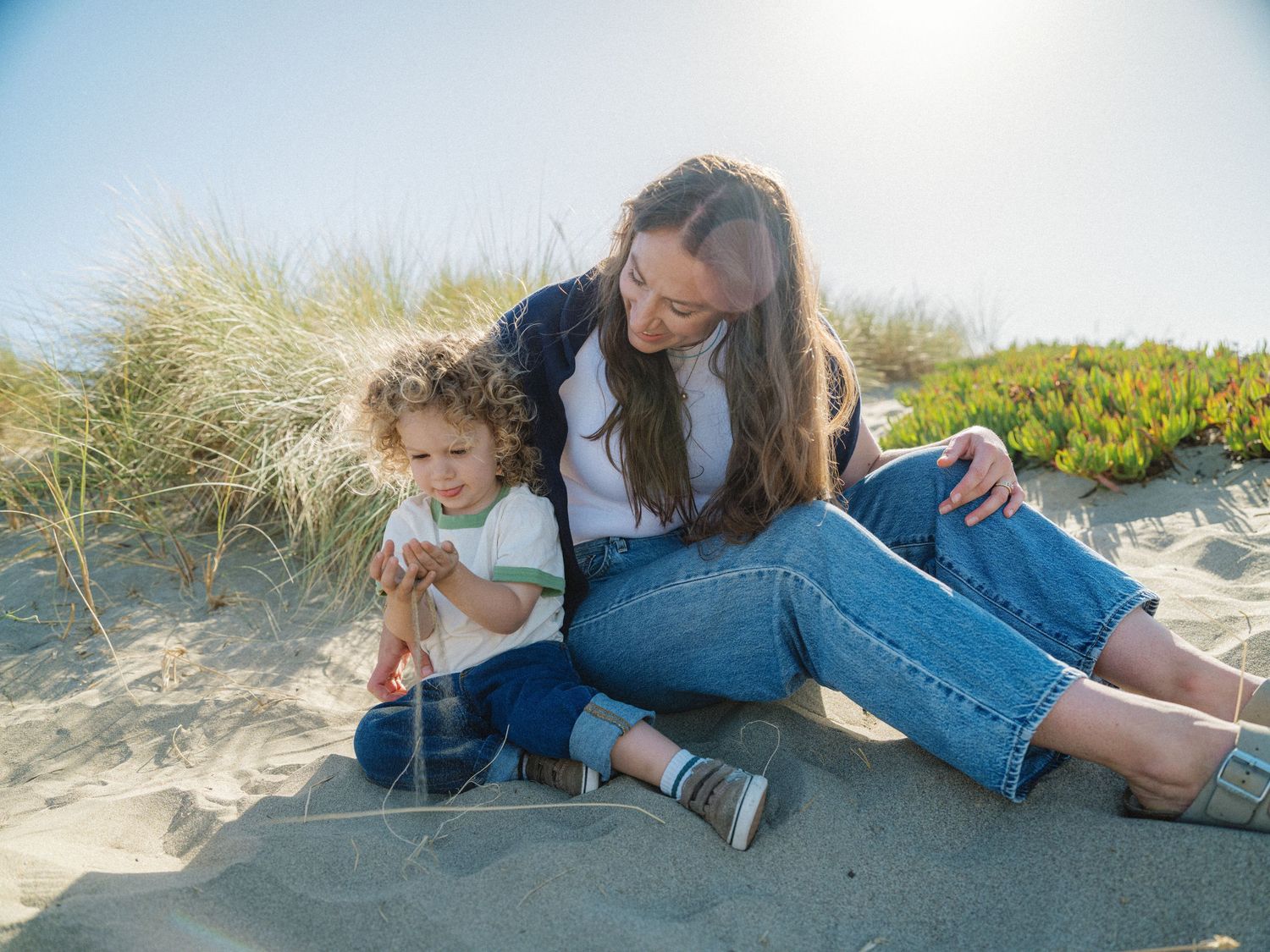 A mother and young child sit together on sandy beach dunes, exploring and playing in the sun.