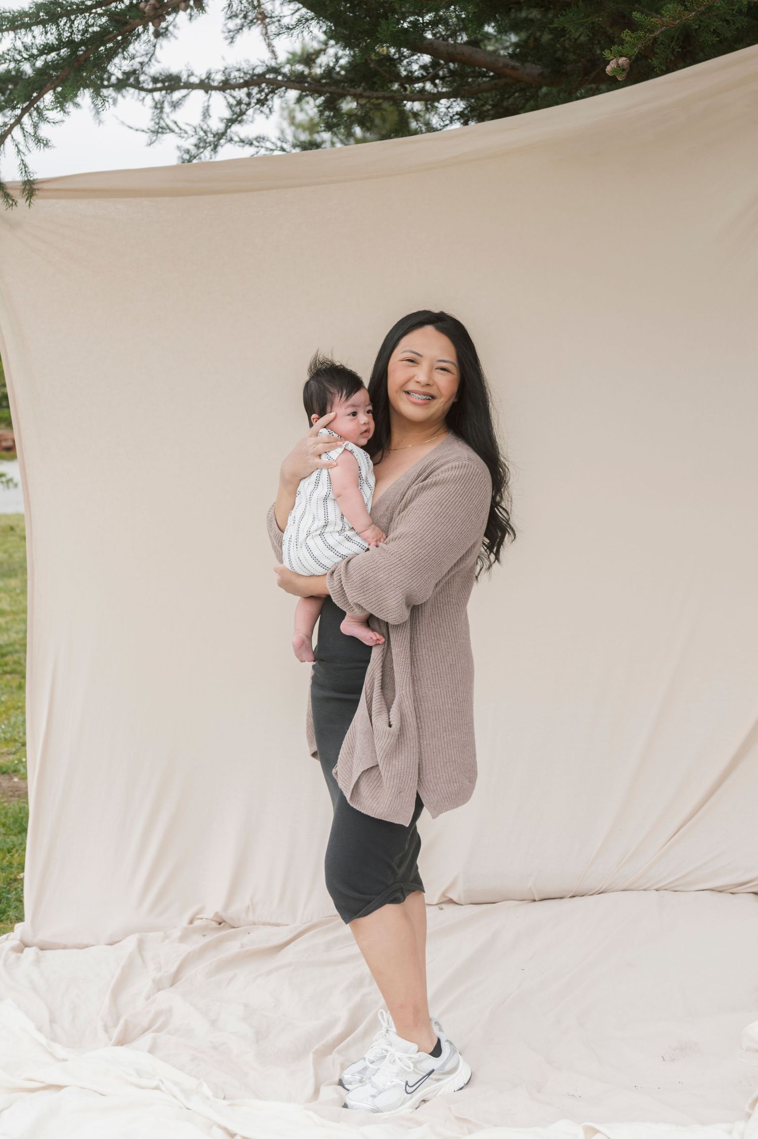 Smiling Asian woman holding a baby in striped outfit, standing against a cream backdrop outdoors.
