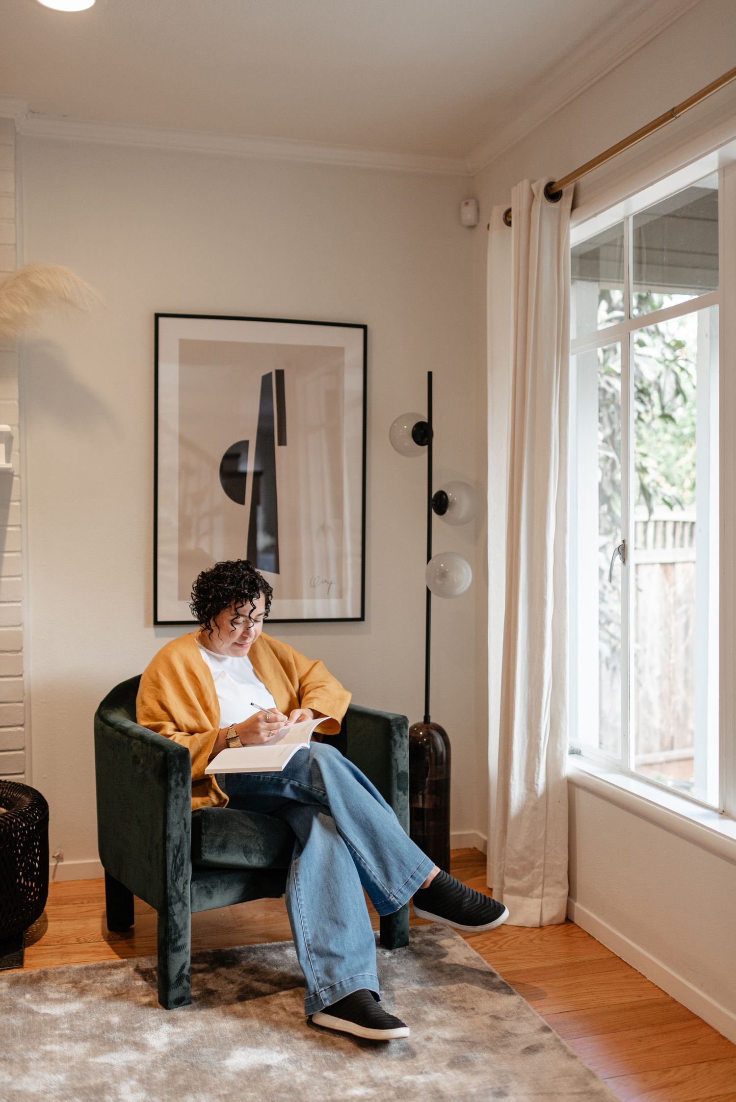 Woman in yellow cardigan and jeans writing in notebook while seated in green velvet armchair by window.