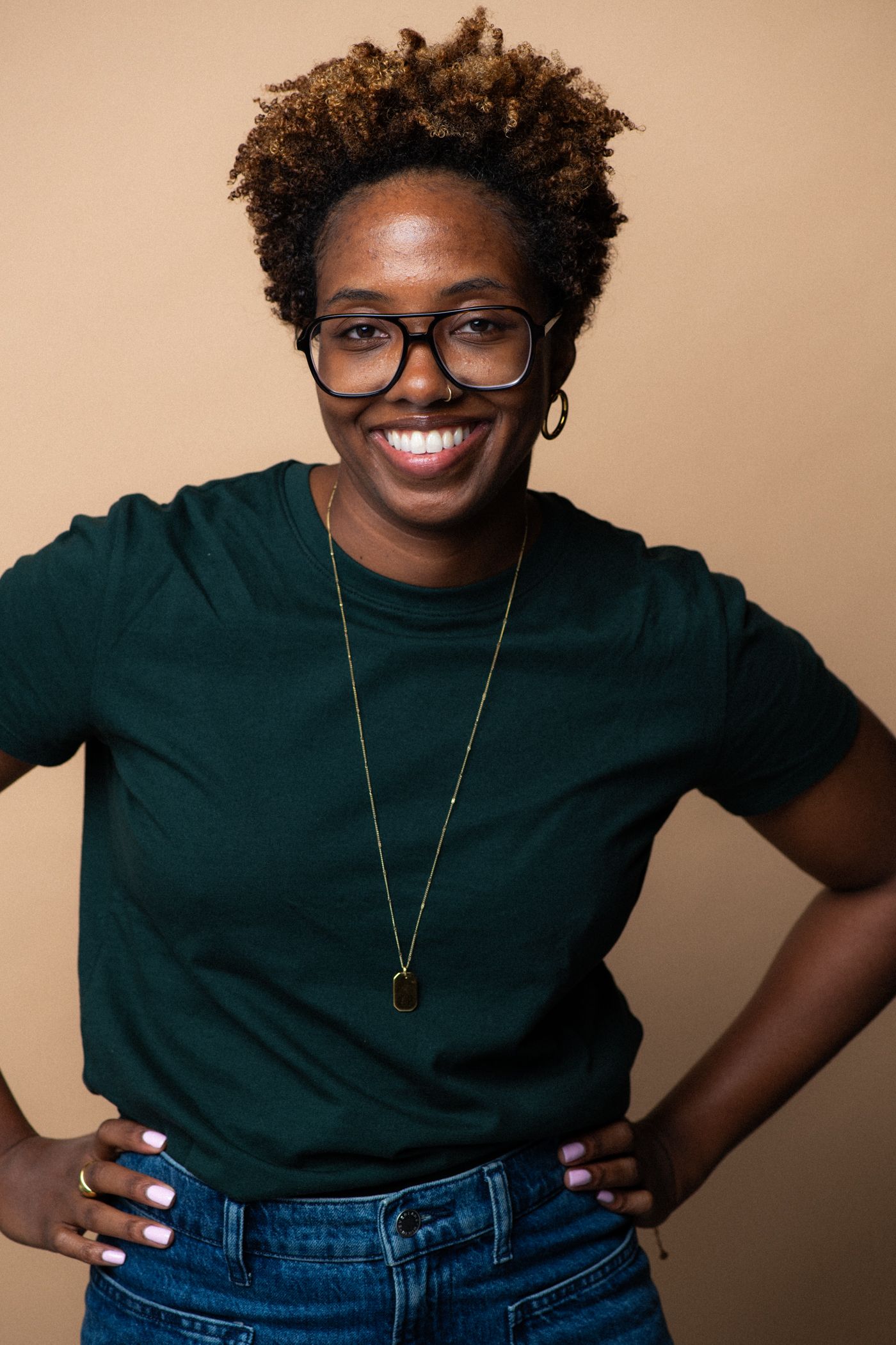 Smiling Black woman with glasses and natural hair wearing a green t-shirt and jeans, posing confidently with hands on hips.