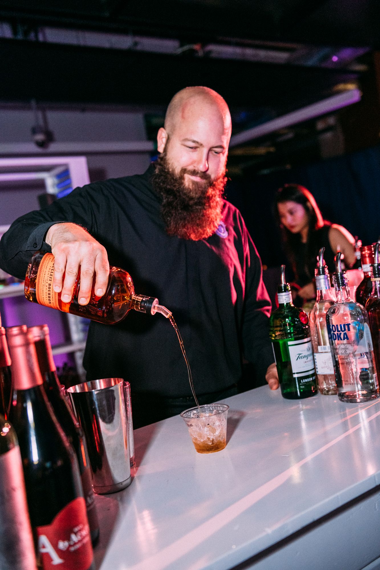 A bearded bartender in black pours multiple cocktails simultaneously at a dimly lit bar counter.