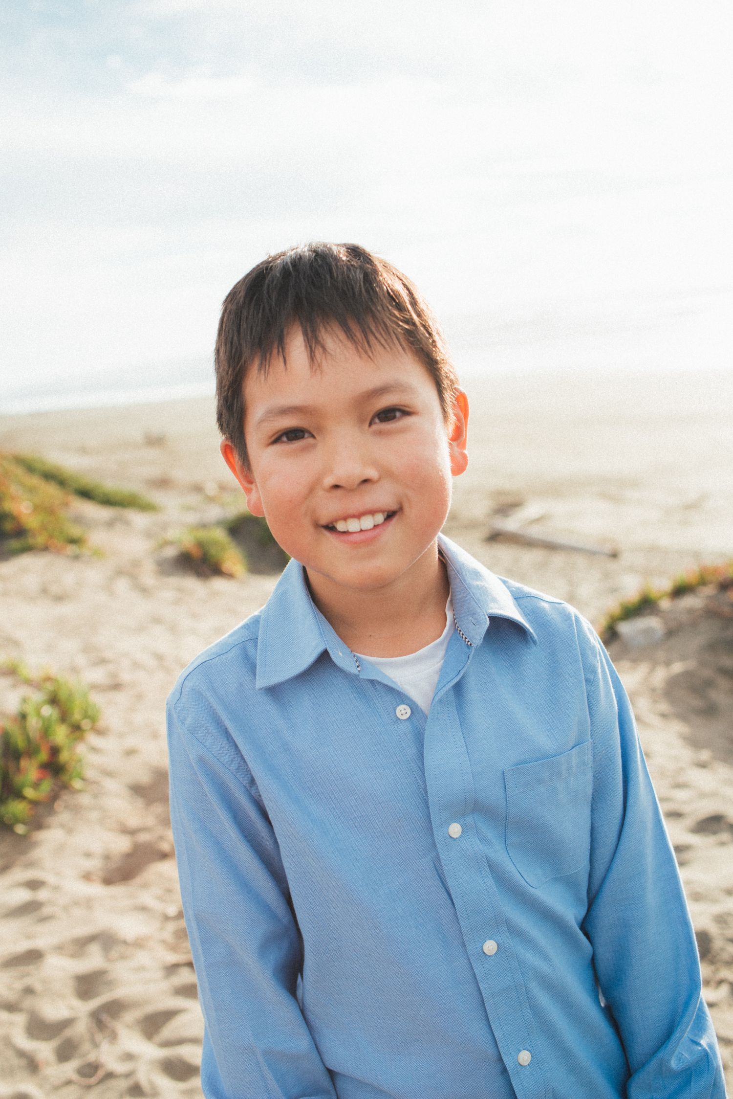 A young boy in a light blue button-down shirt smiles at the beach.