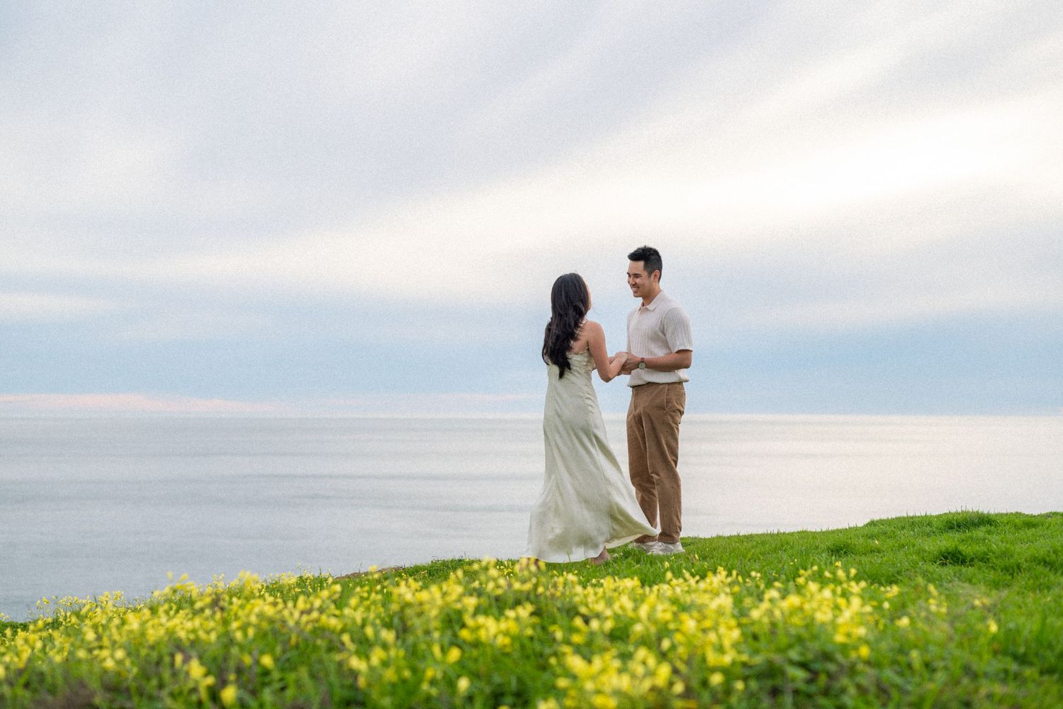 A couple stands together in a scenic field of yellow wildflowers overlooking the ocean on a cloudy day.