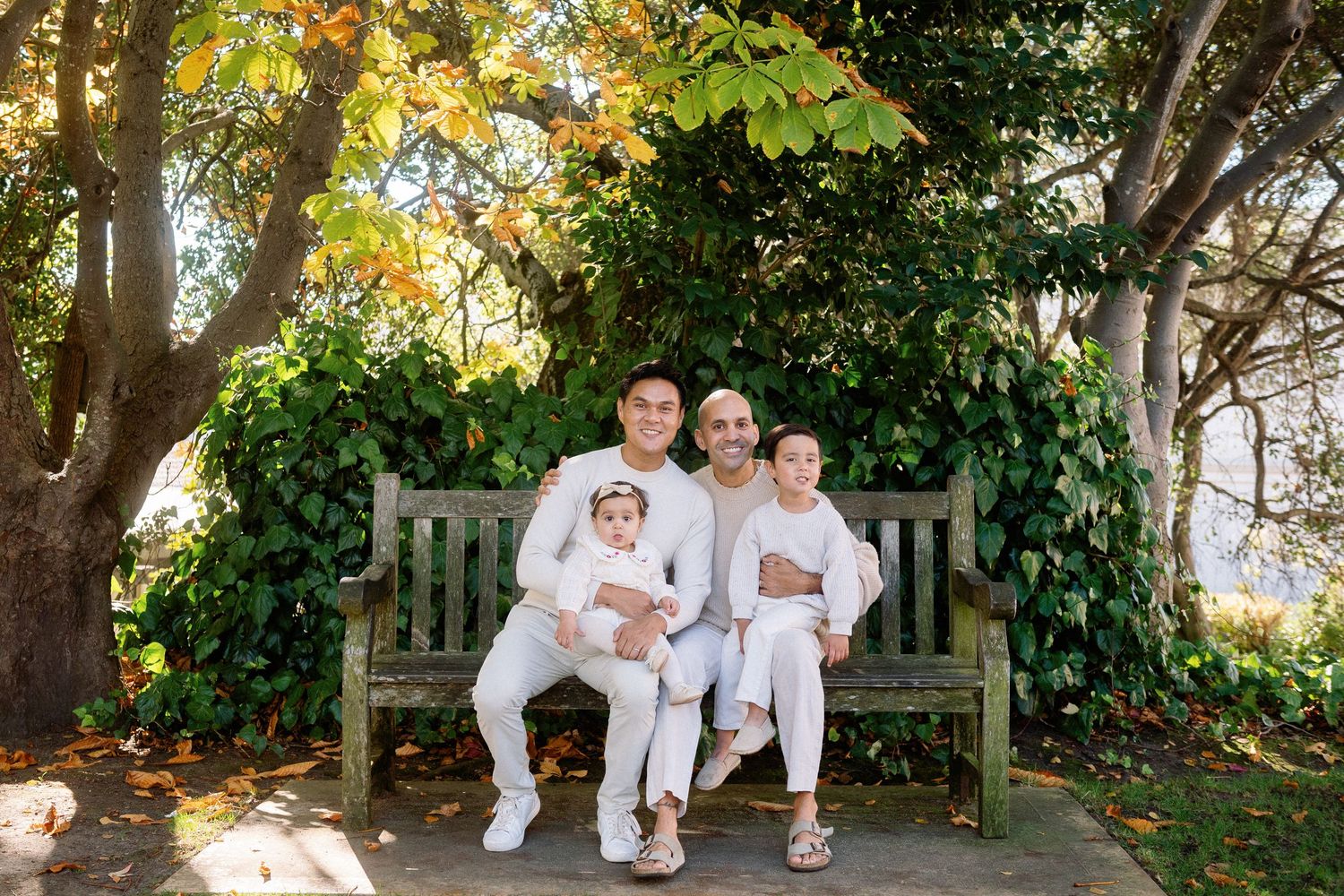 Family dressed in white poses together on wooden bench in garden setting.