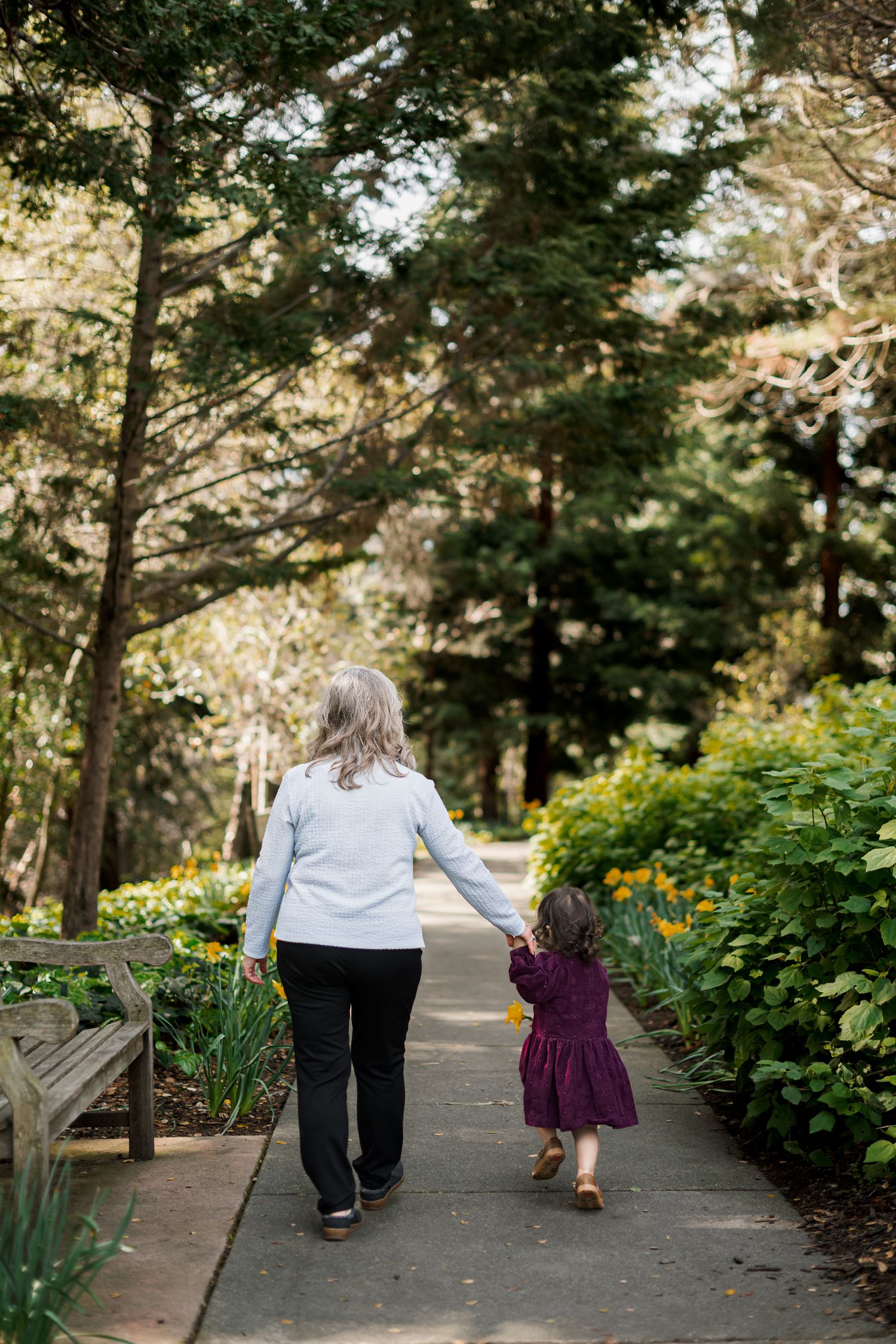 Grandmother and granddaughter walking hand in hand along a garden path surrounded by trees and flowers.