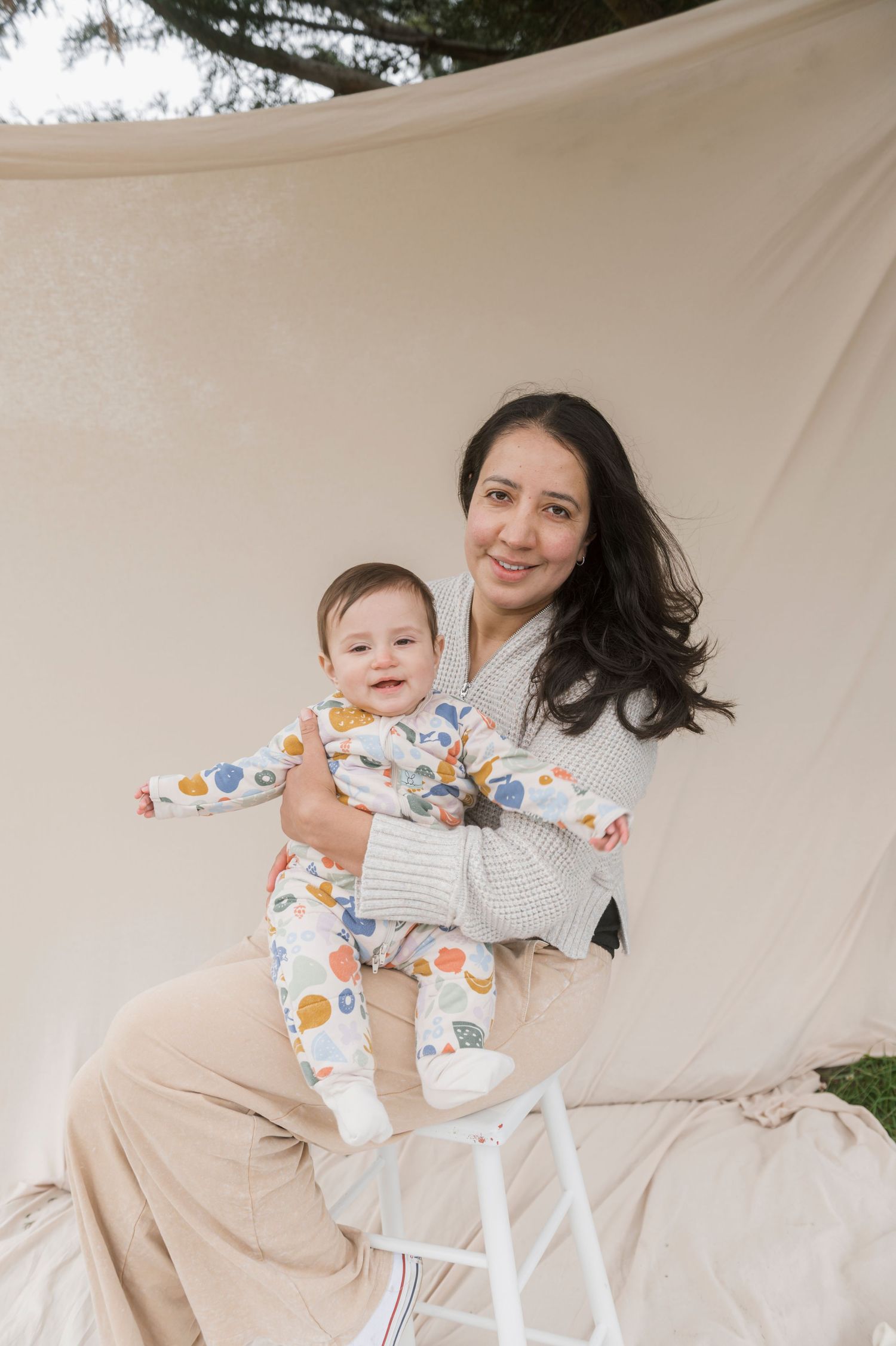 Smiling mother holding baby in colorful printed onesie, seated on stool against cream fabric backdrop outdoors.