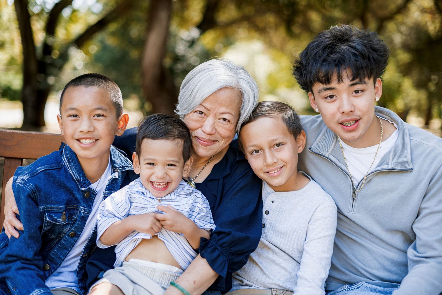 Grandmother sitting on a bench in a park smiling with four grandchildren of mixed ethnicities.
