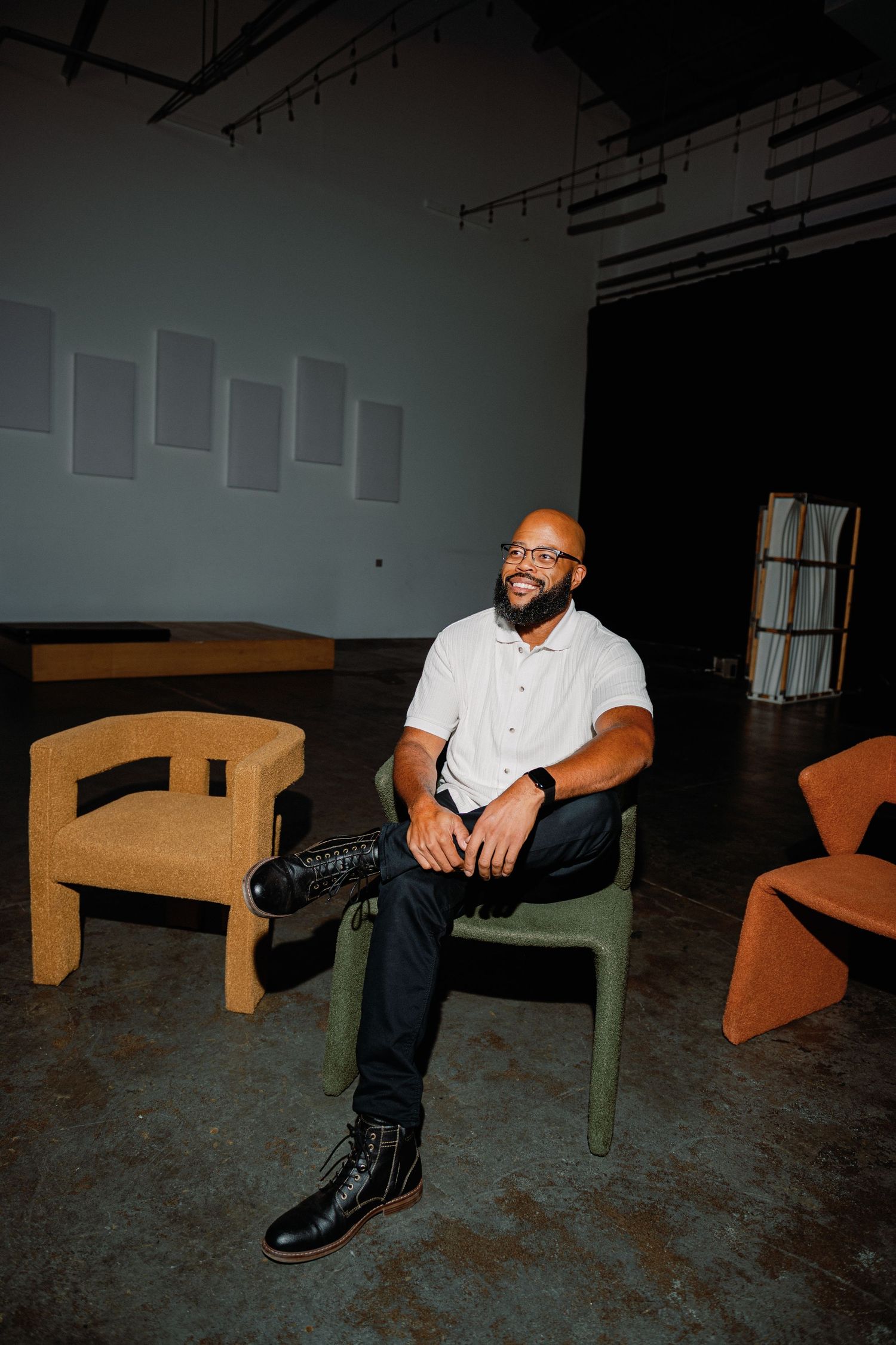 A bald man with glasses sits smiling in a green chair inside a modern art gallery with cork furniture.