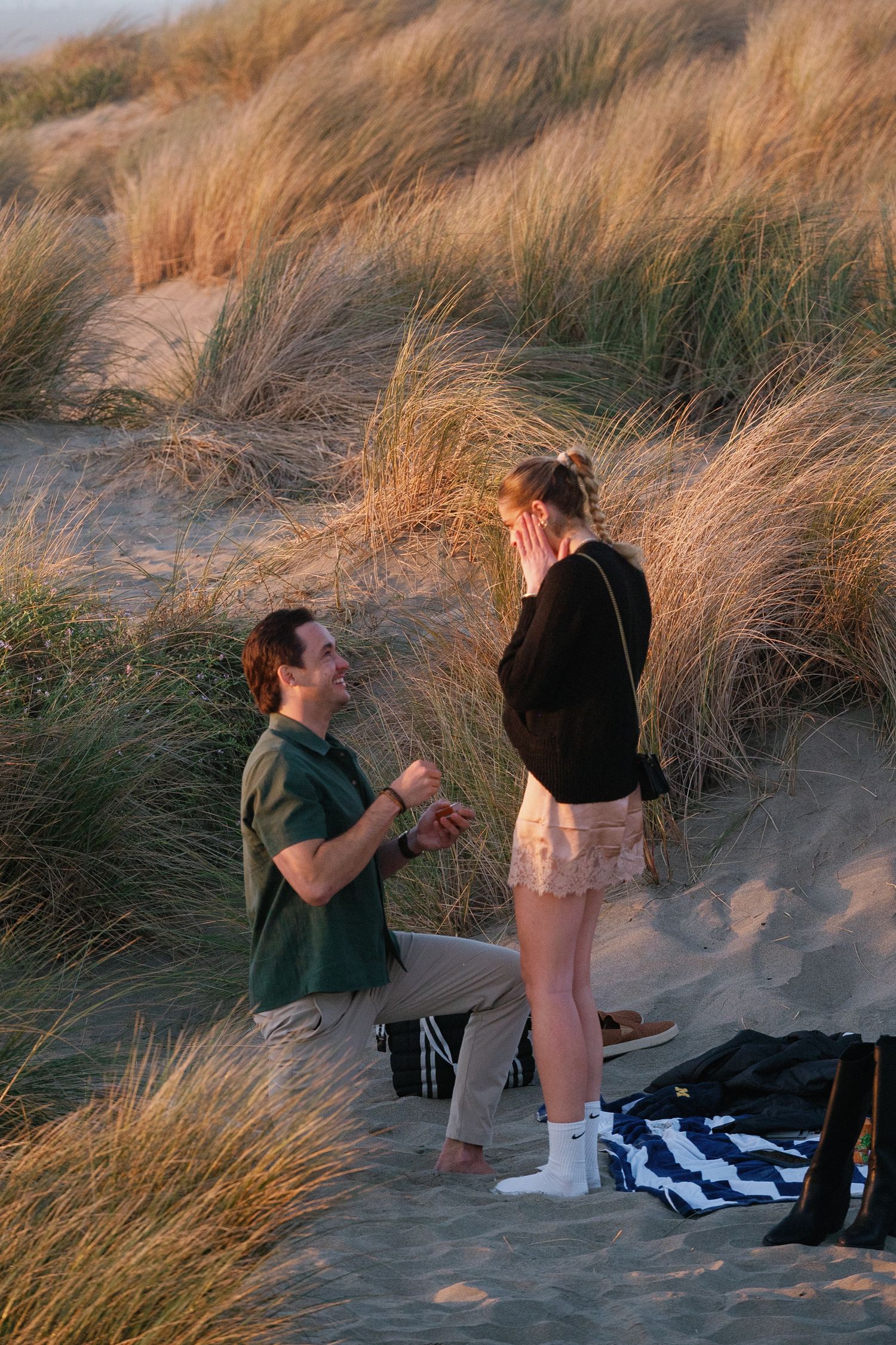 Surprise proposal moment captured on beach with tall grass during sunset.