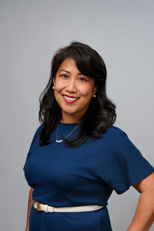 Professional woman in navy blue dress with pearl necklace smiling confidently against gray background.