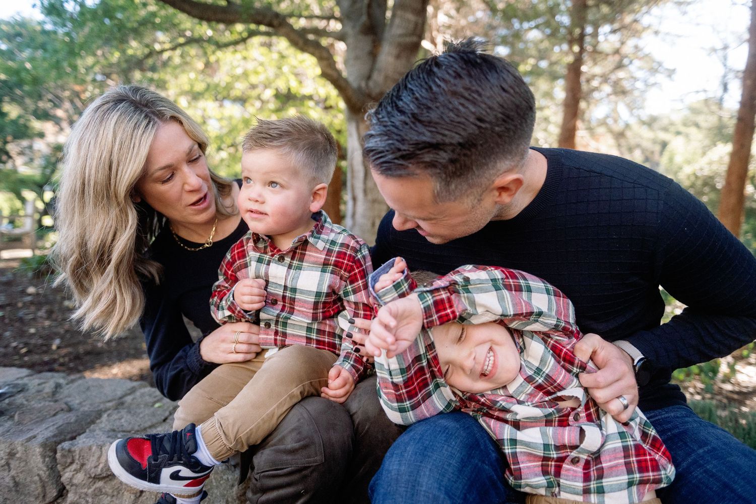 Family of four in matching plaid outfits posing outdoors among trees, parents smiling at two young children.