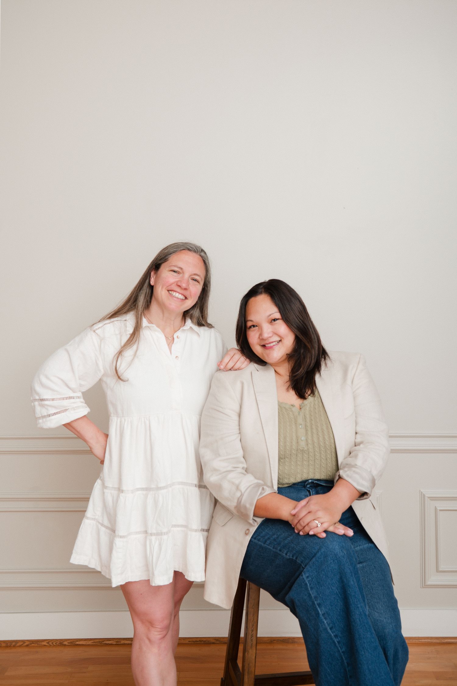Two women smiling together in a bright studio, one in a white dress and one in a blazer with jeans.