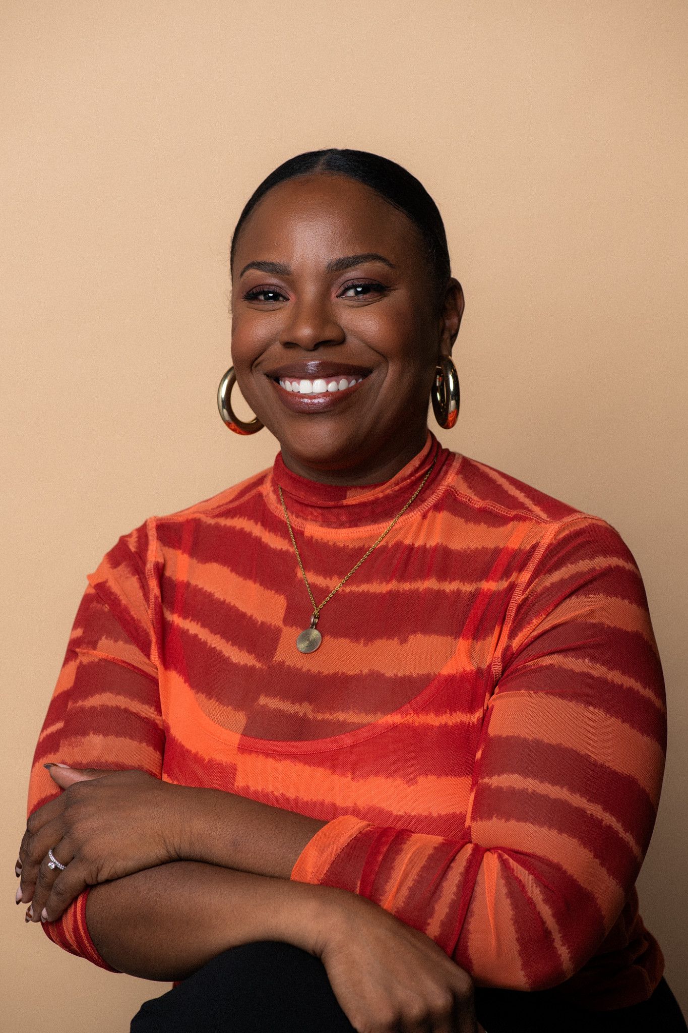Smiling woman in red striped turtleneck top with hoop earrings and green pendant necklace, arms crossed, beige background.