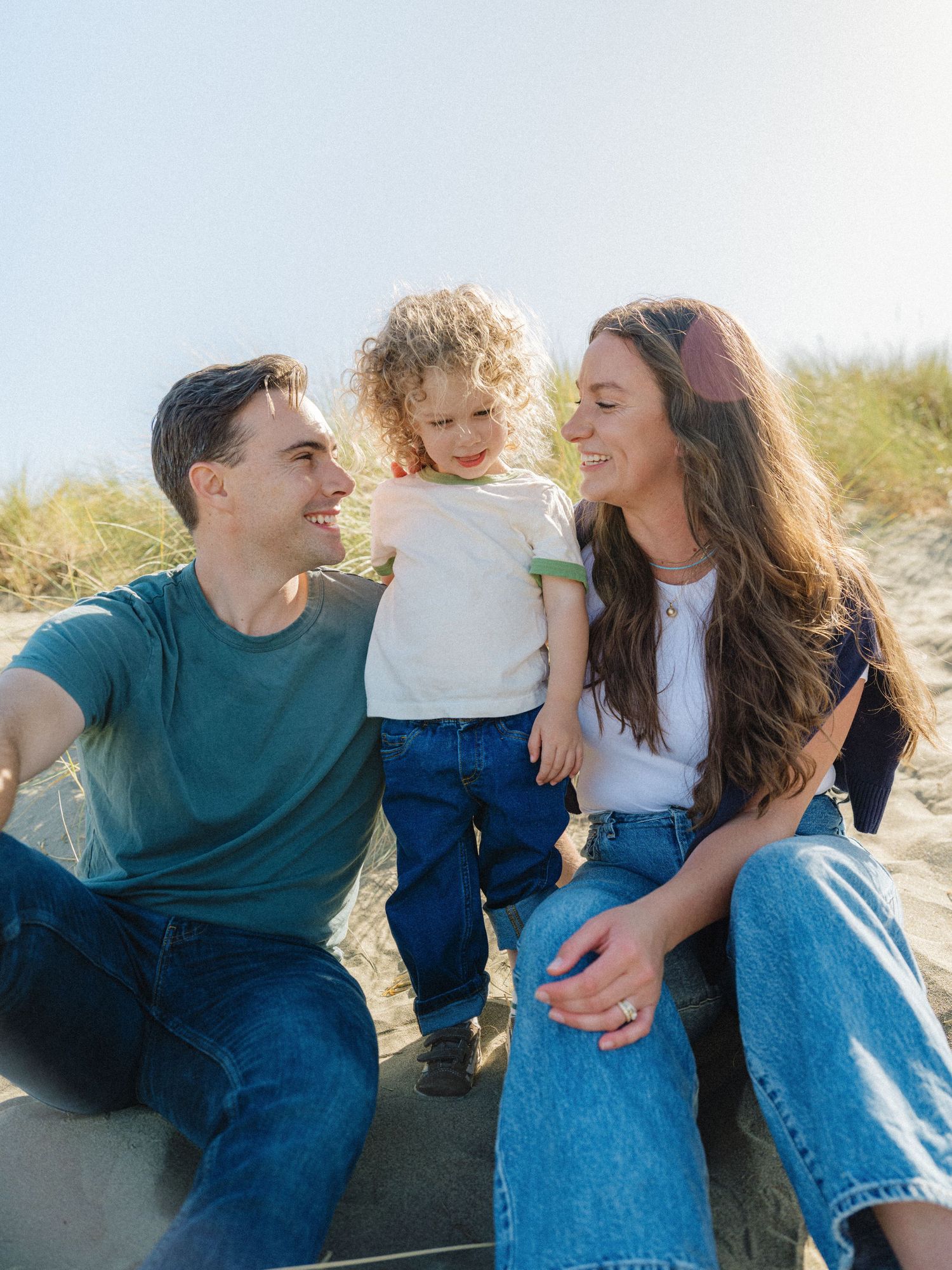 A happy young family with a curly-haired toddler sits together on sandy beach dunes on a sunny day.