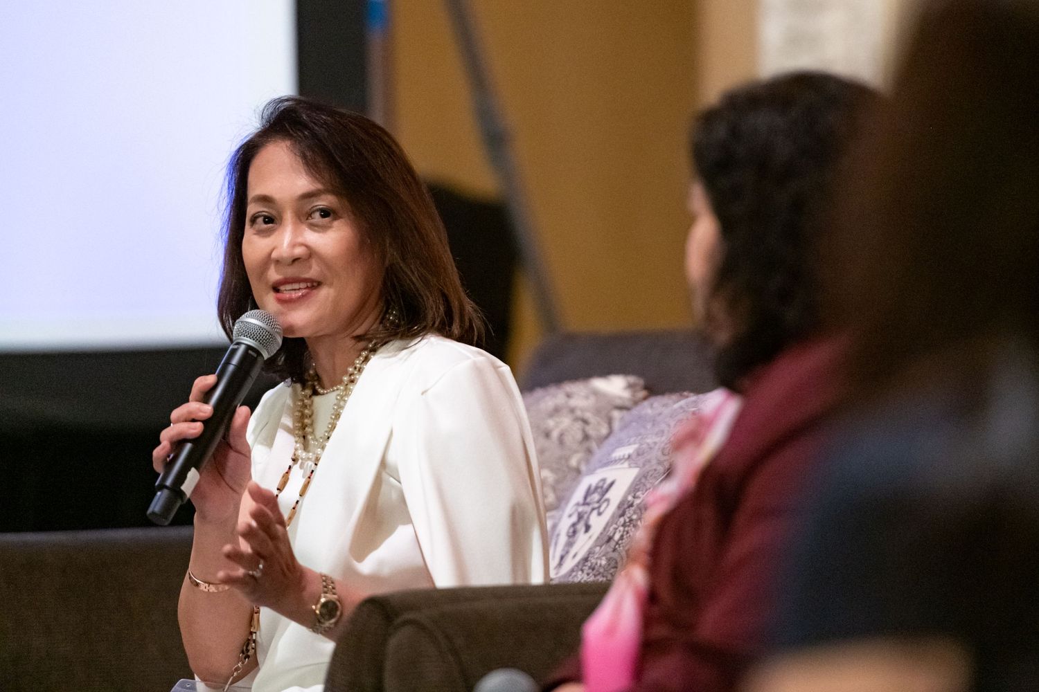 Woman in white outfit speaking at panel discussion, holding microphone, engaged in conversation at professional event.