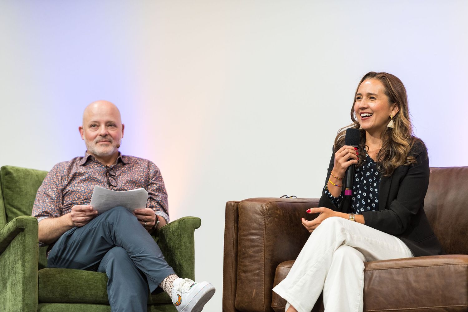 Two professionals sit in armchairs during a panel discussion, man holds tablet, woman speaks into microphone.