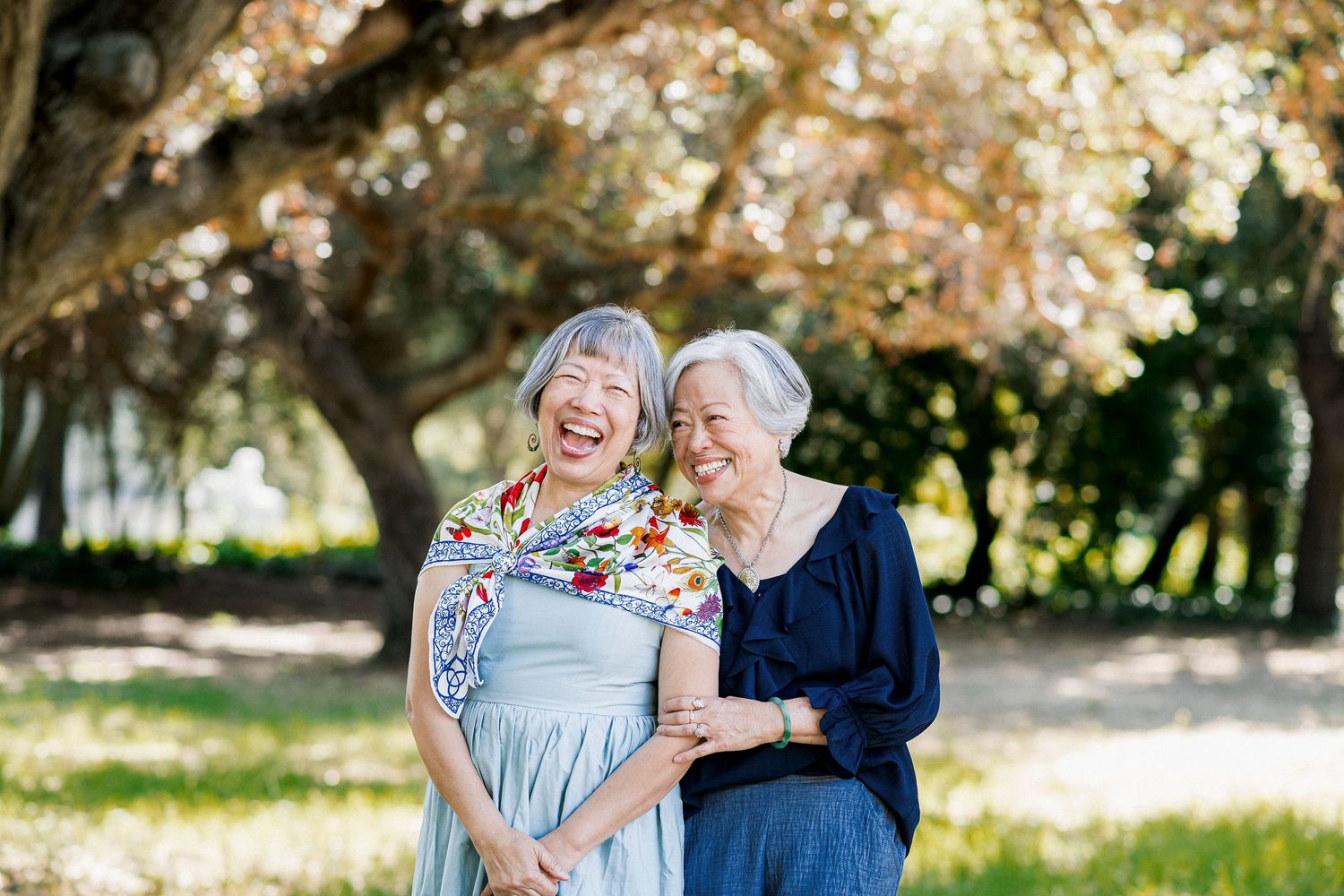 Two elderly Asian women laughing joyfully together outdoors in a sunny park with trees in the background.