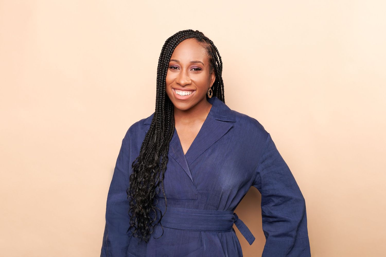 Smiling woman in navy blue wrap dress with long braids poses confidently against a beige background.