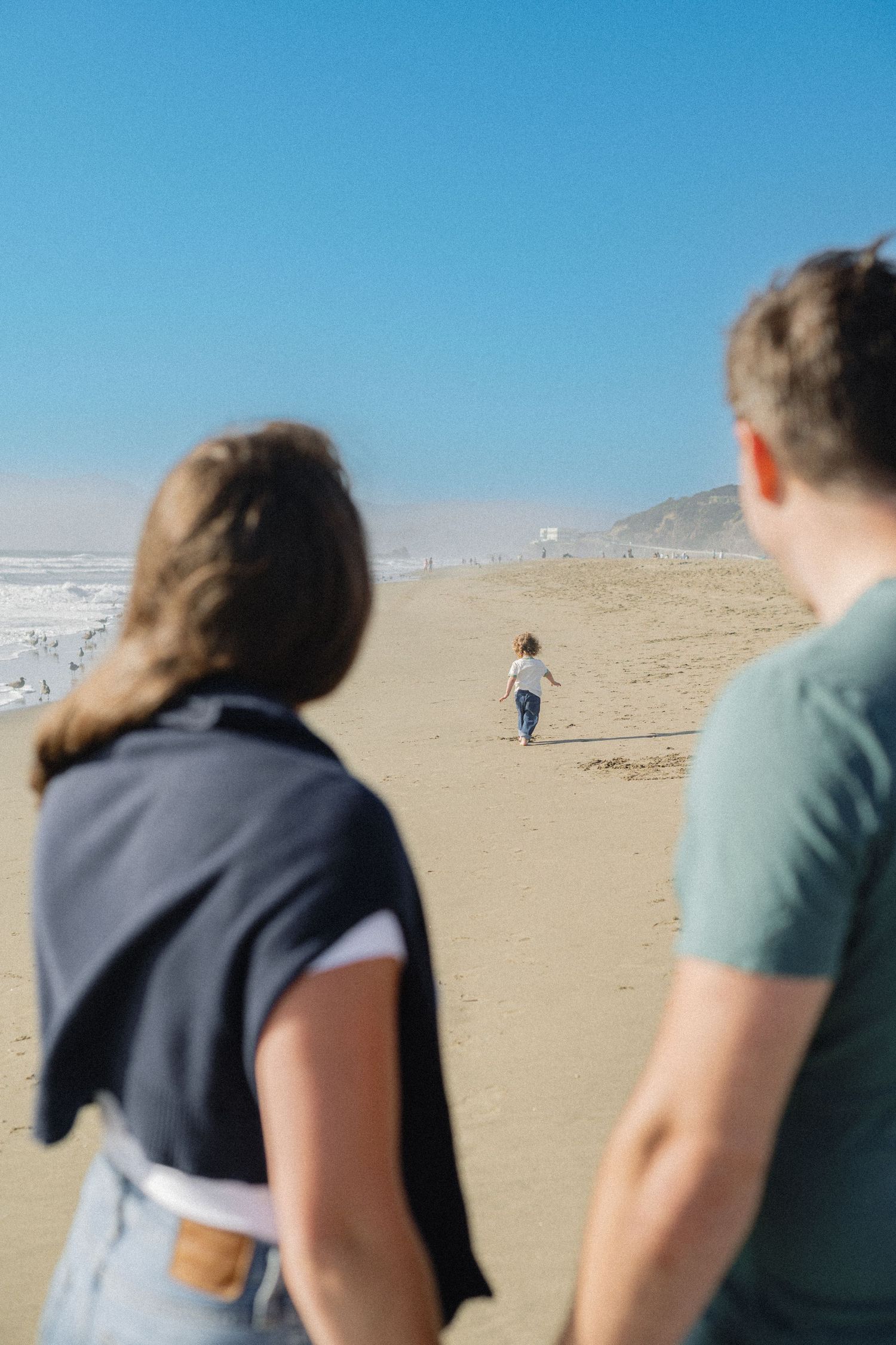 Couple watching their young child run across a sandy beach under a clear blue sky.