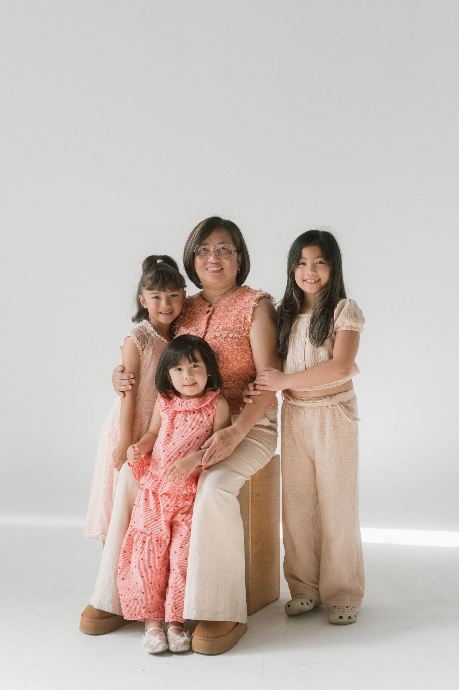 A grandmother poses with three young girls in coordinating neutral and pink outfits against a white studio background.