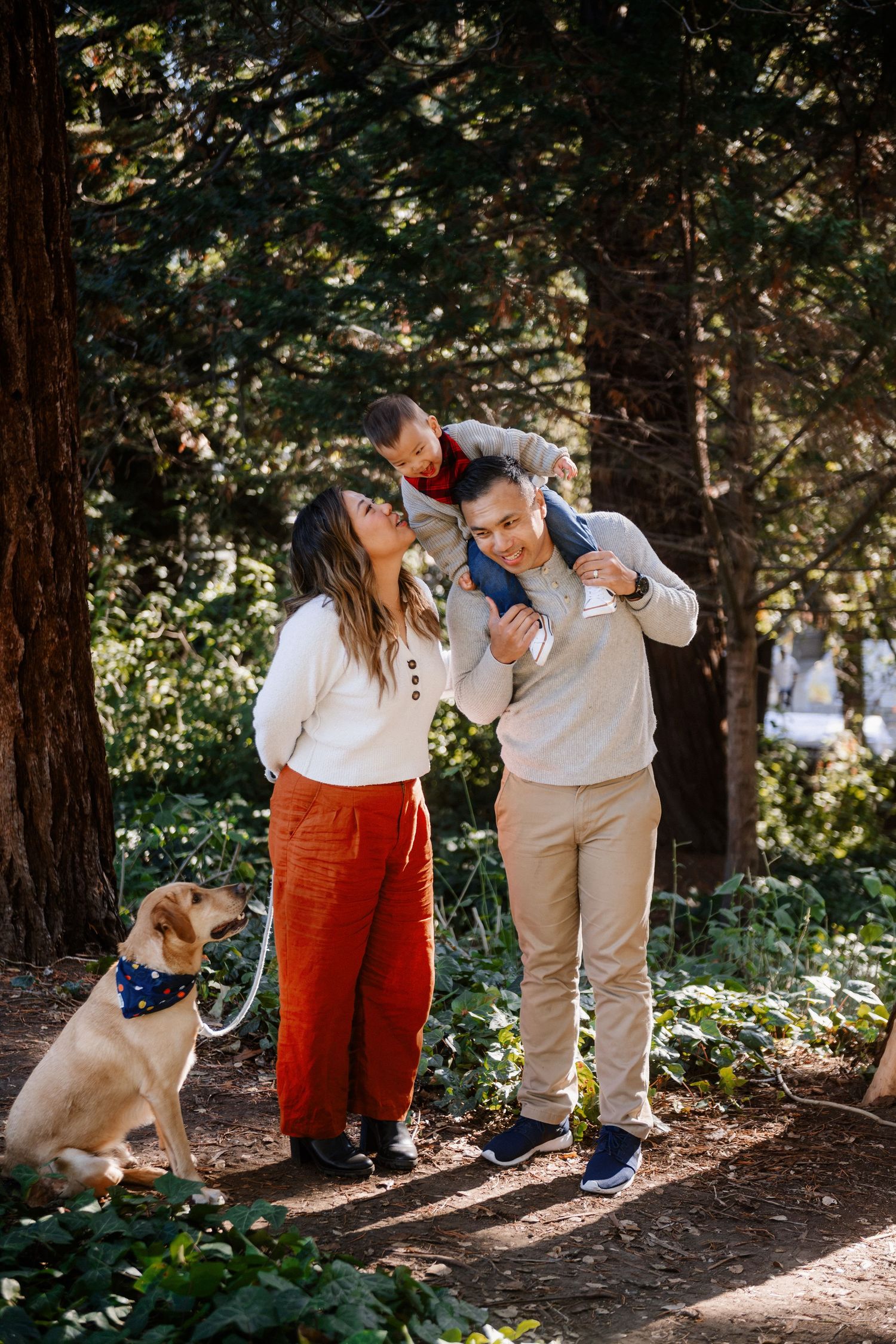 Family with young child on dad's shoulders and golden dog on leash smiling together in sunlit forest.