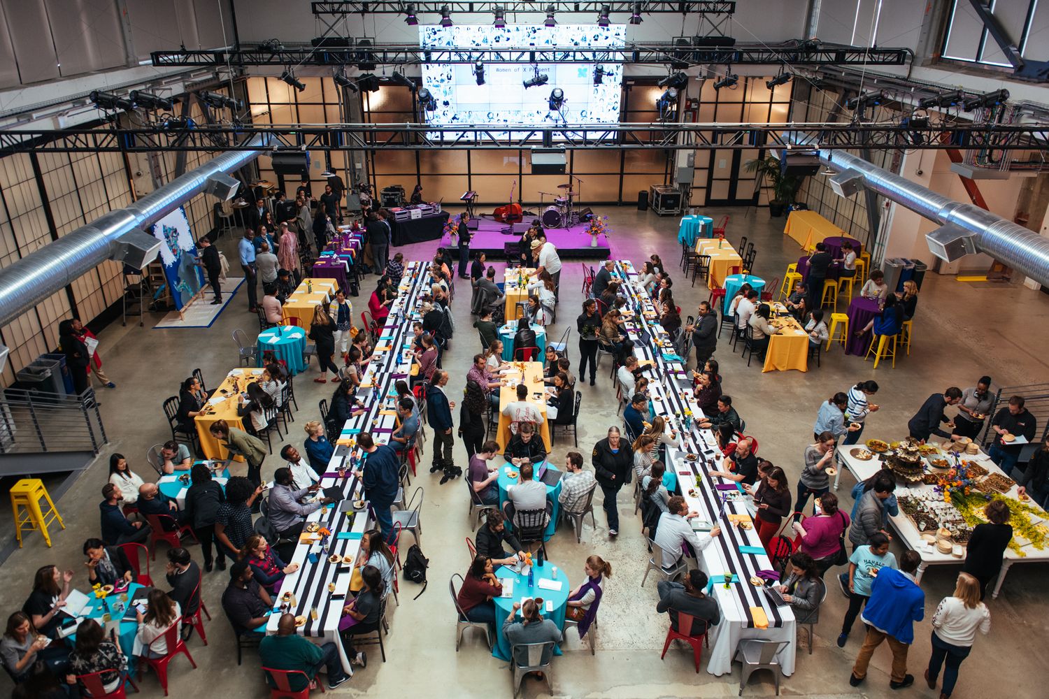 Large group of people seated at long tables in a cafeteria-style space during a community gathering event.