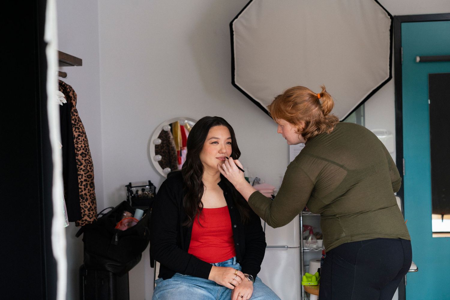 Makeup artist applying cosmetics to a young woman seated in a studio with professional lighting equipment.