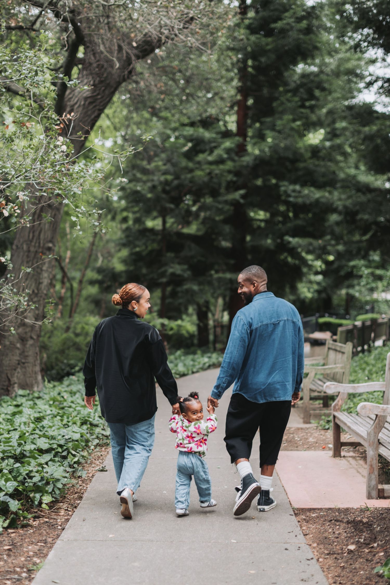 A family of three walks hand-in-hand down a park path, parents holding their toddler between them surrounded by lush green trees.