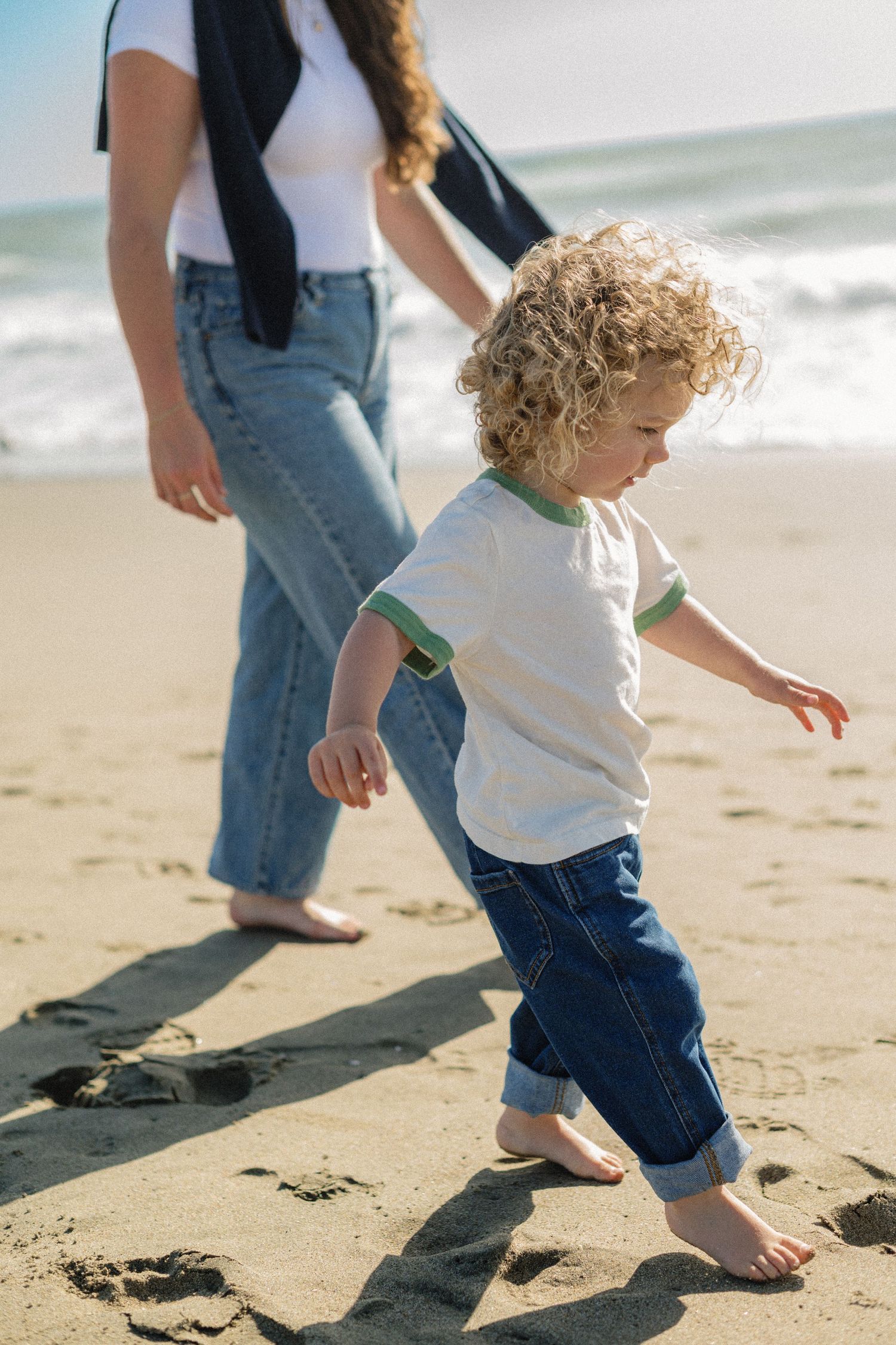 Curly-haired toddler walking barefoot on sandy beach with mother in jeans and white top near ocean waves.