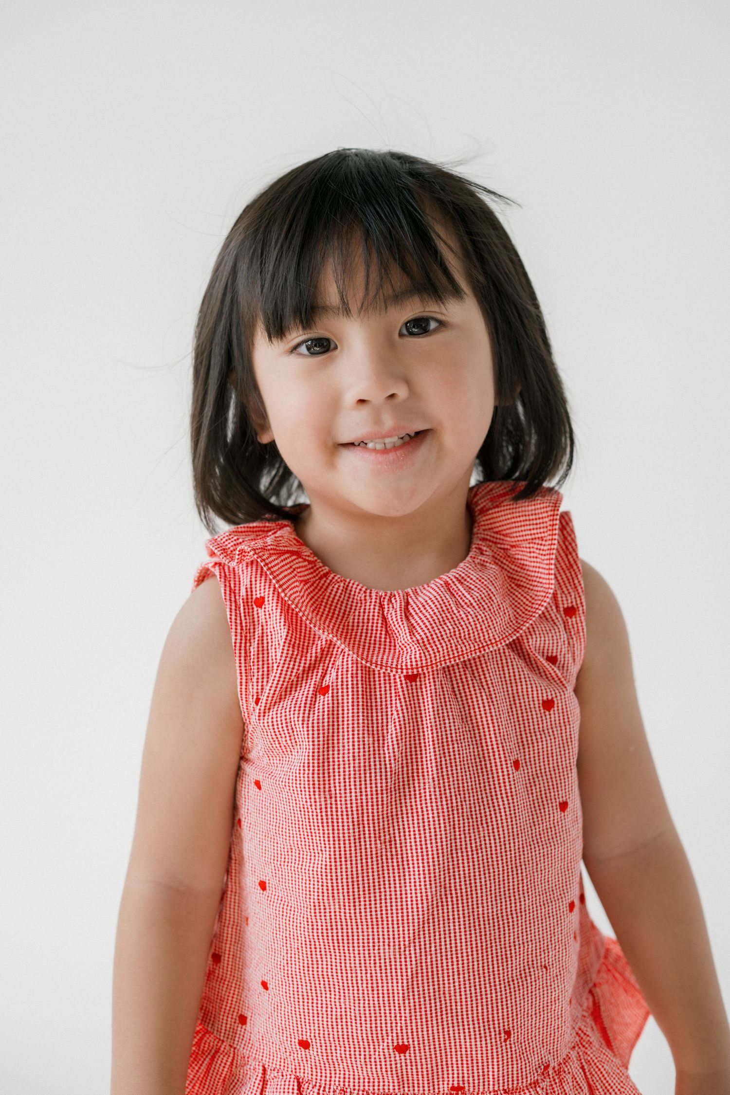 A smiling young Asian girl with black hair wearing a red striped sleeveless dress with ruffle collar on white background.