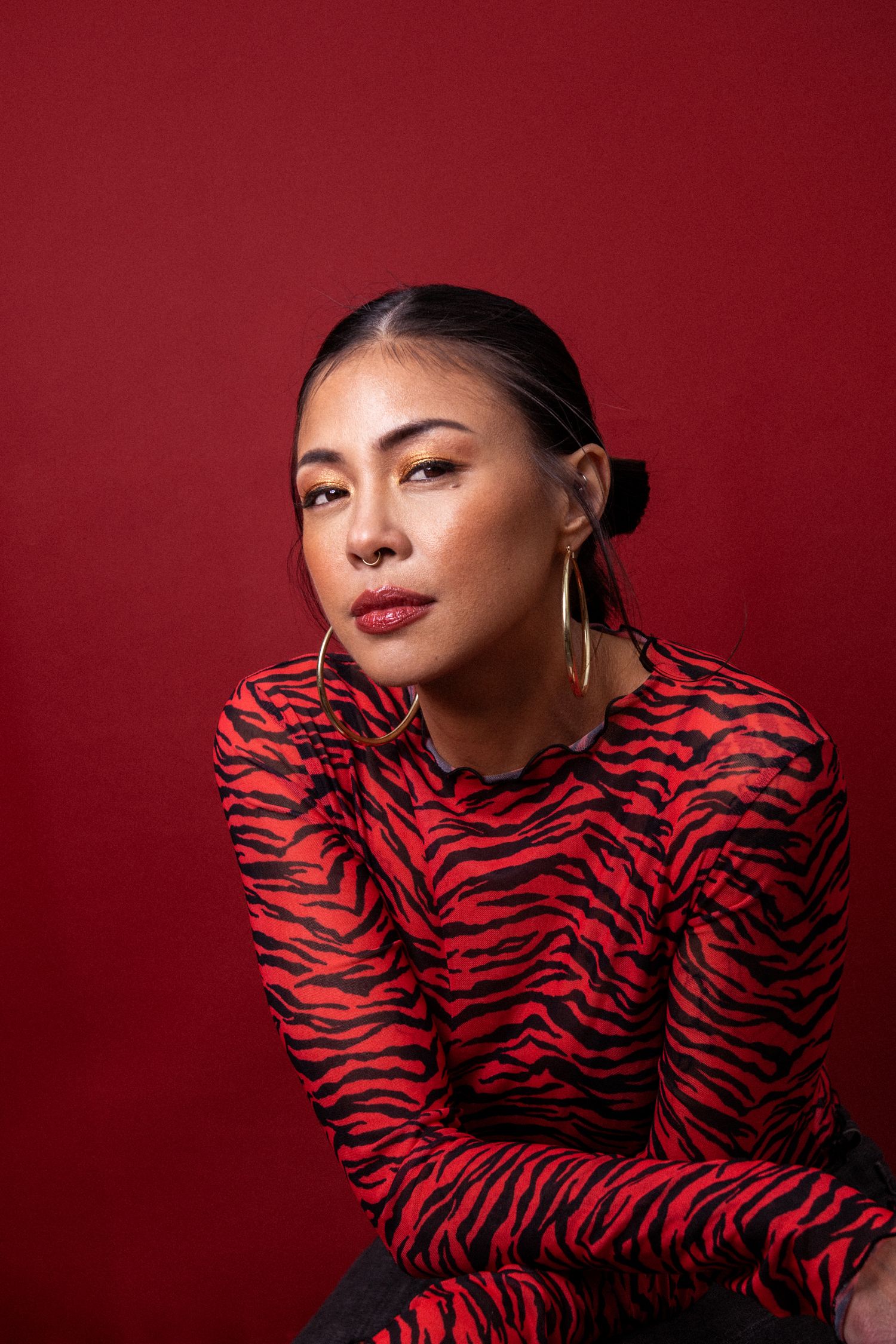 Woman in red tiger-print top with gold hoop earrings poses against a bold red background.