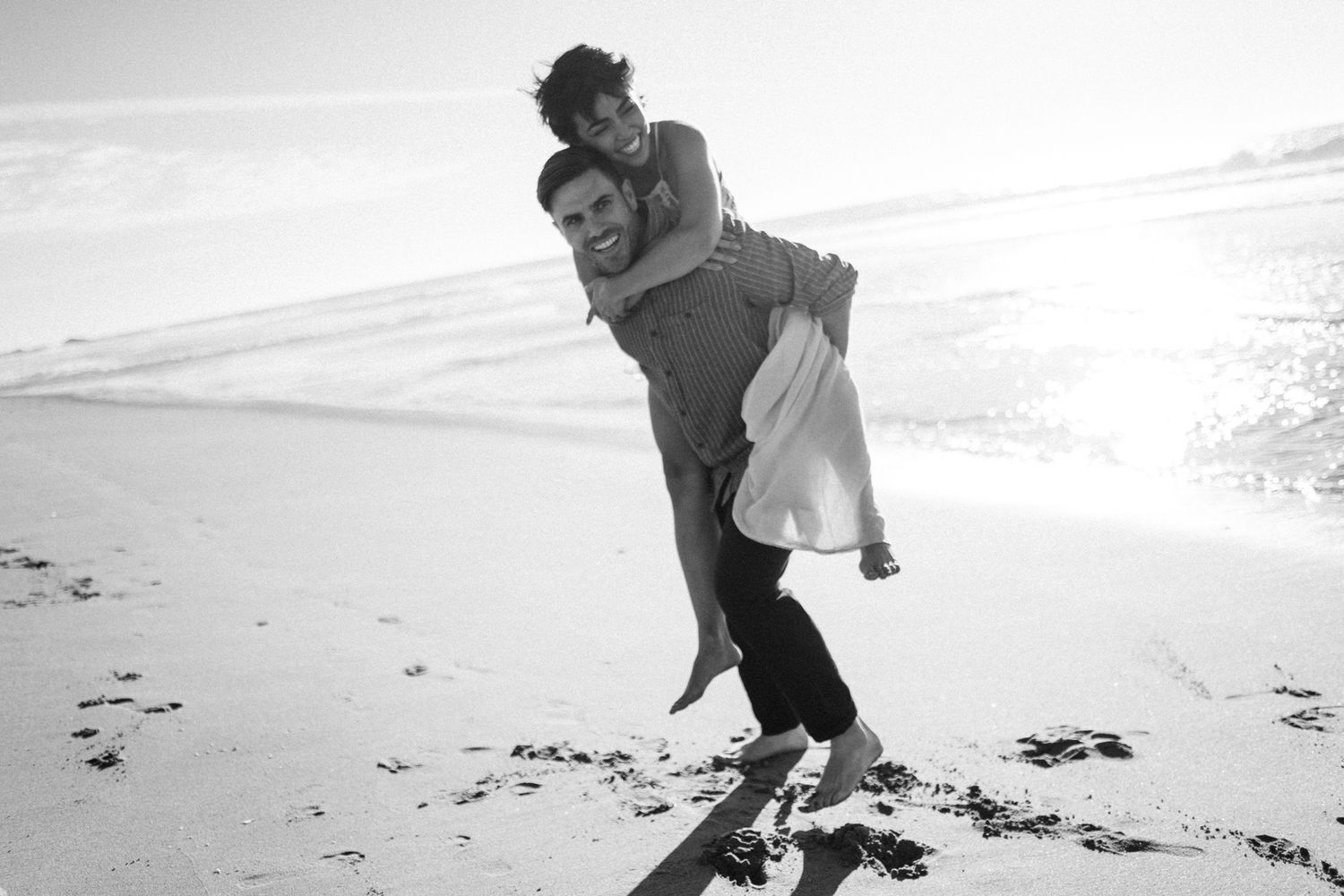 A couple embraces on a beach in black and white as waves crash behind them creating a romantic seaside moment.