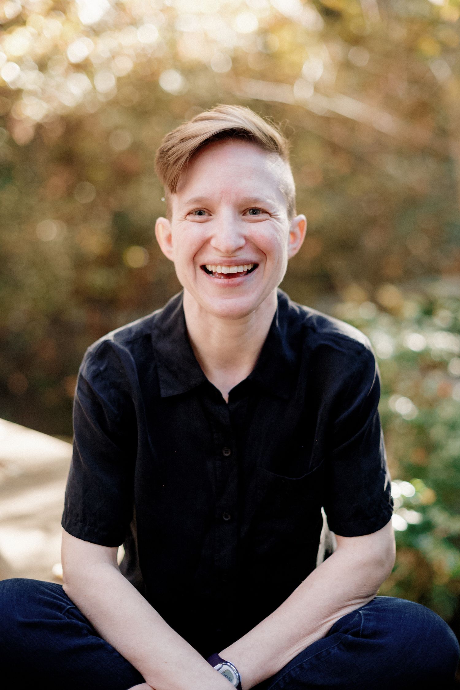 Person in black polo shirt sitting outdoors with natural bokeh background.