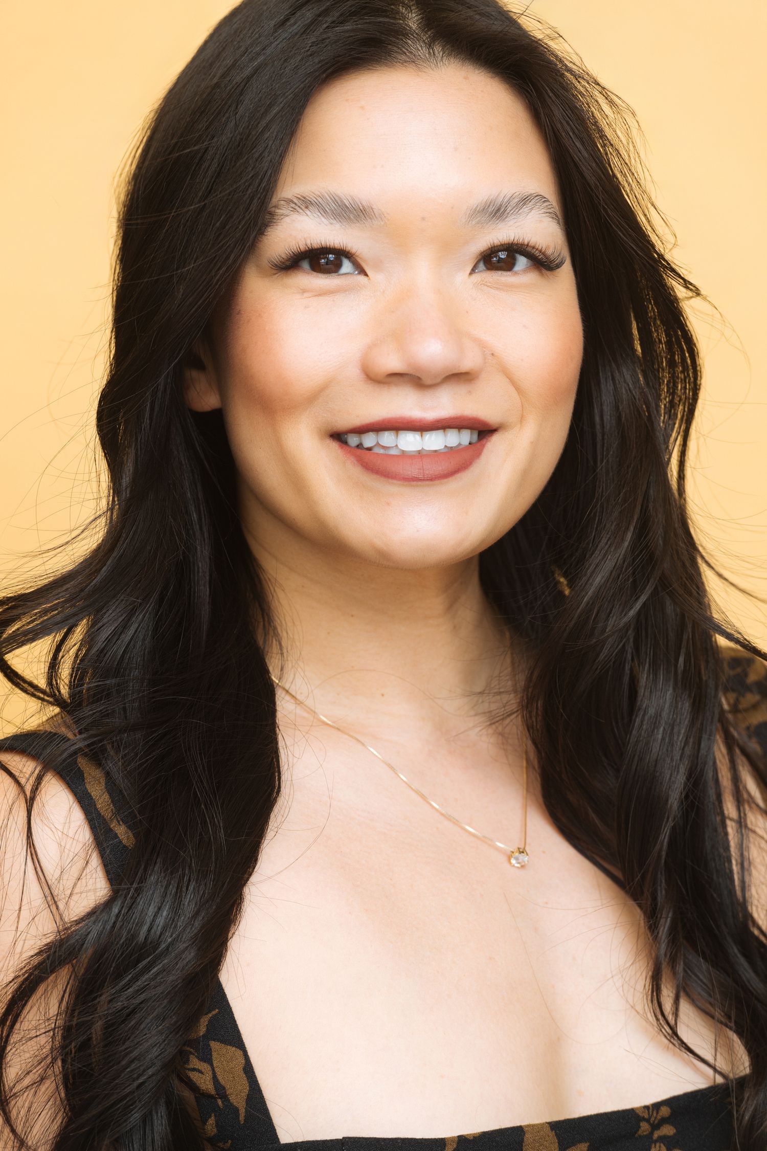 Smiling Asian woman with long black hair wearing a black top and gold necklace against a yellow background.
