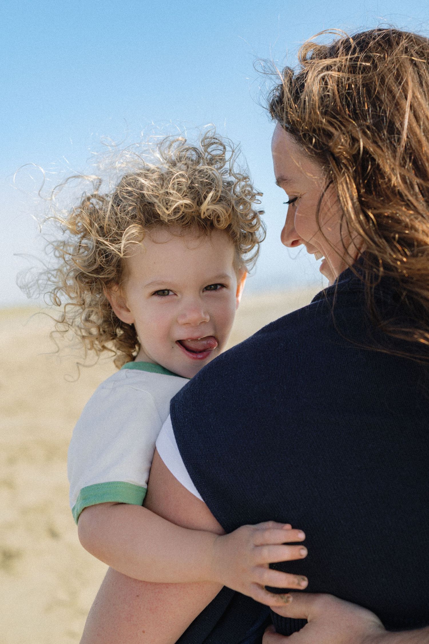 Curly-haired toddler sticking tongue out while being held by mother on a sunny beach.