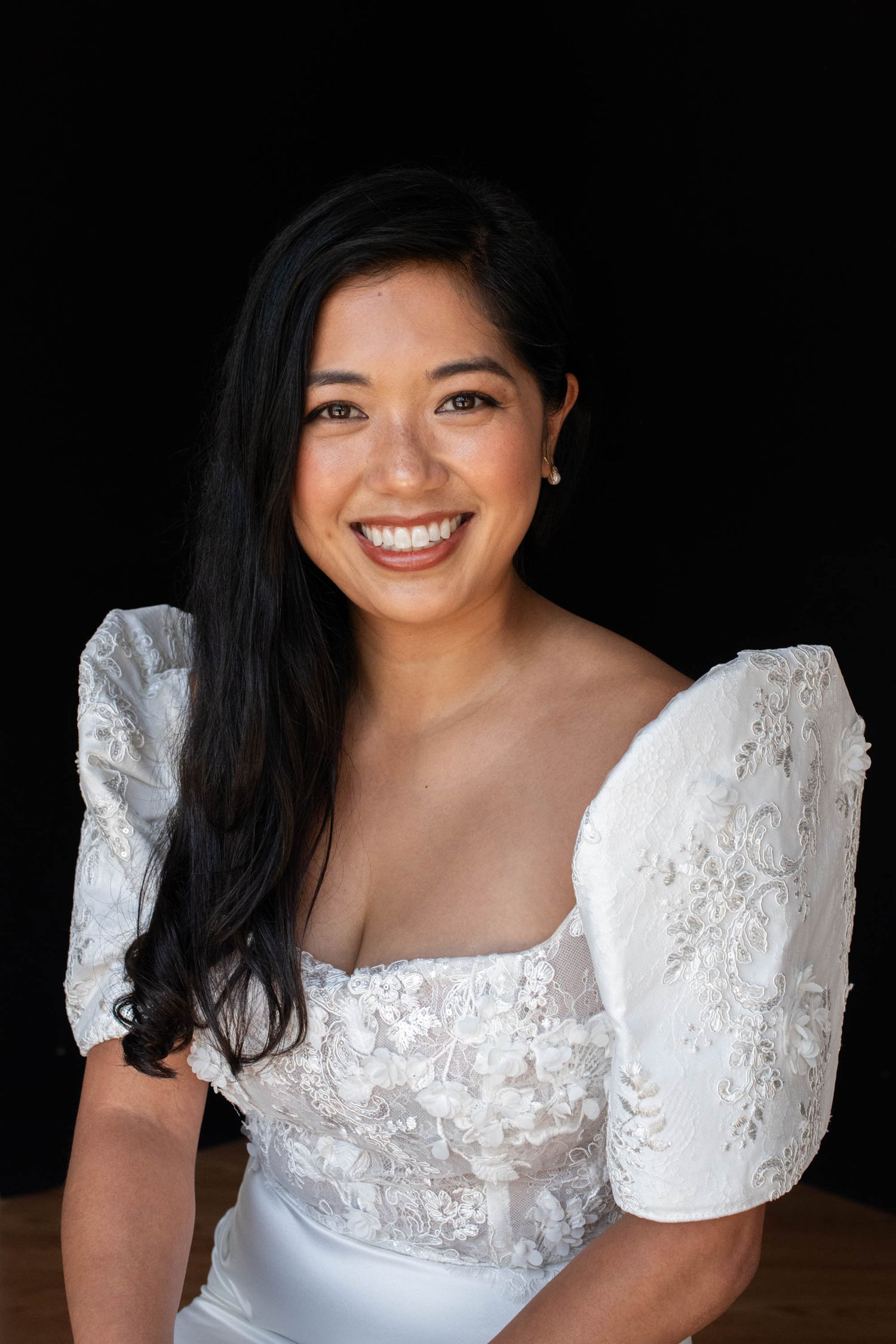 Smiling young woman with long dark hair wearing a white lace puff-sleeve dress against a black background.
