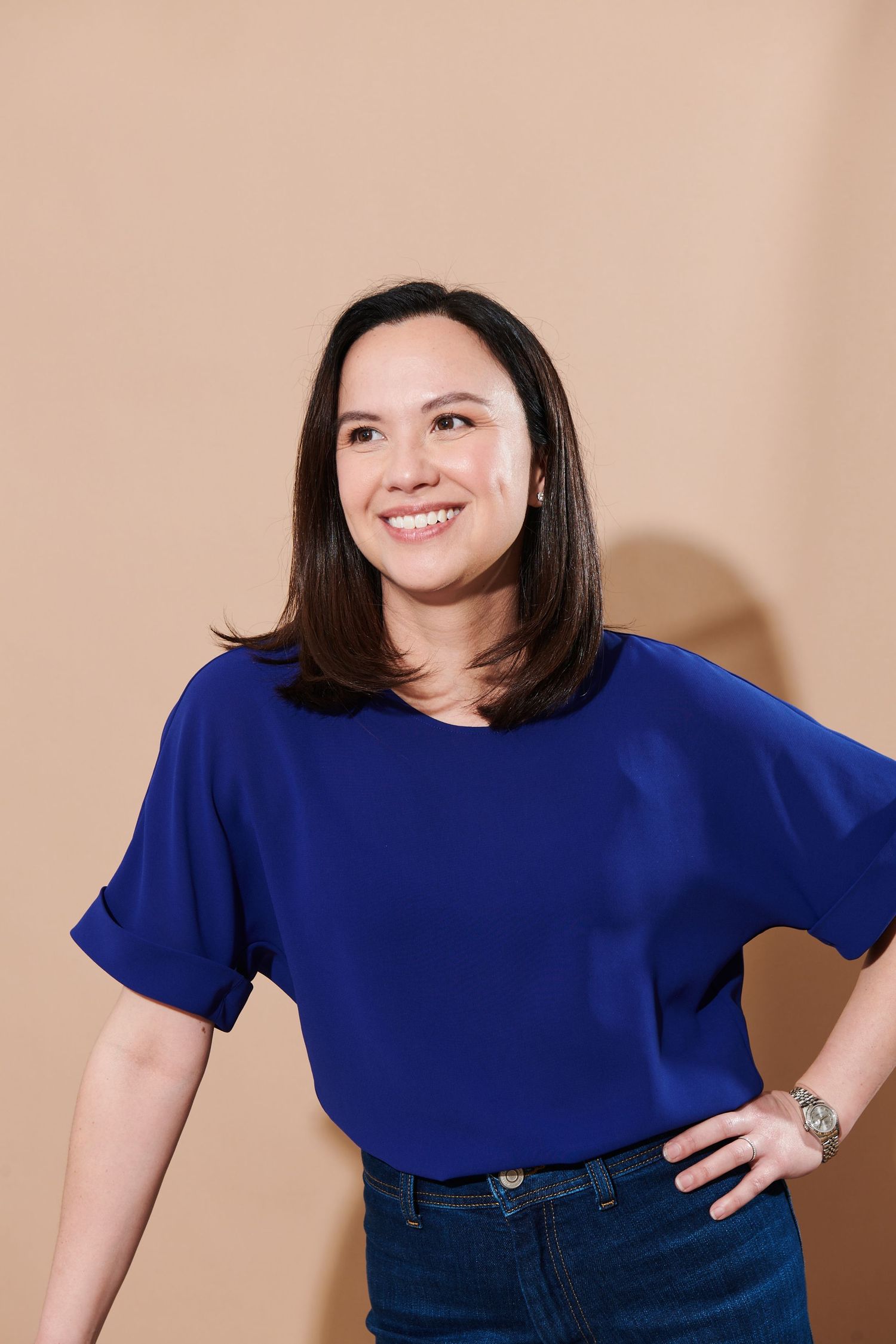 Smiling woman in blue top and jeans posing confidently against a beige background.