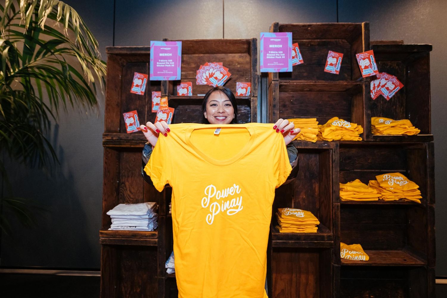 Woman holding yellow Power of Journey t-shirt in front of wooden shelves stocked with merchandise.